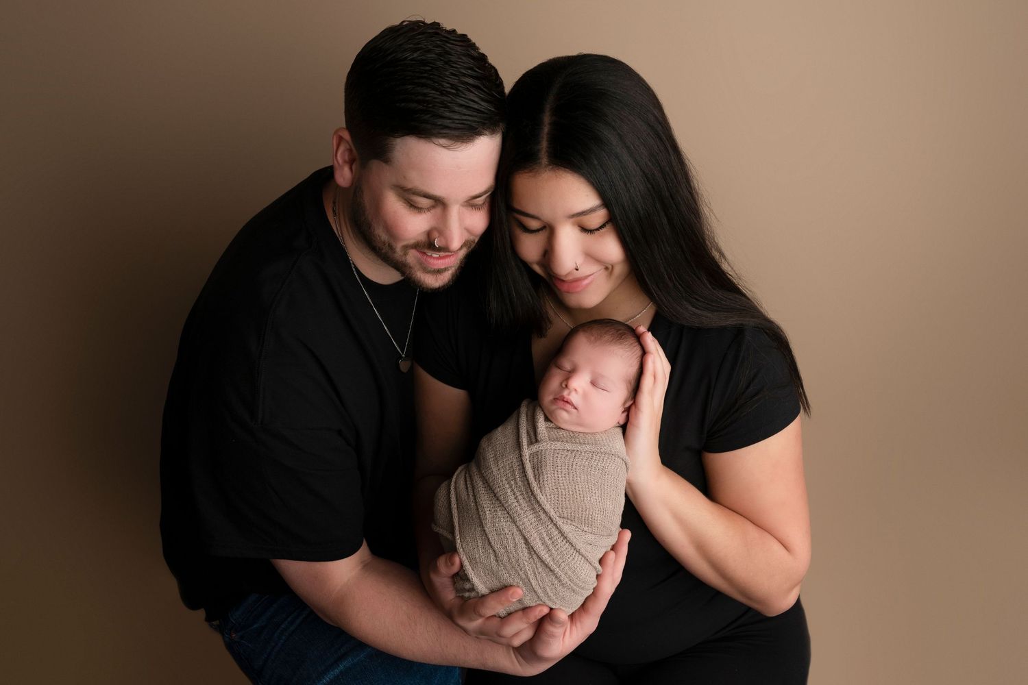 Parents in black shirts tenderly hold their sleeping newborn wrapped in beige, posed against a warm tan studio backdrop.