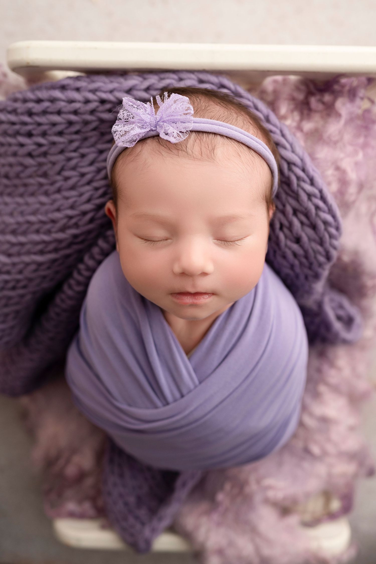 Sleeping newborn baby girl swaddled in purple wrap with floral headband, posed in a knit blanket for a studio photo.