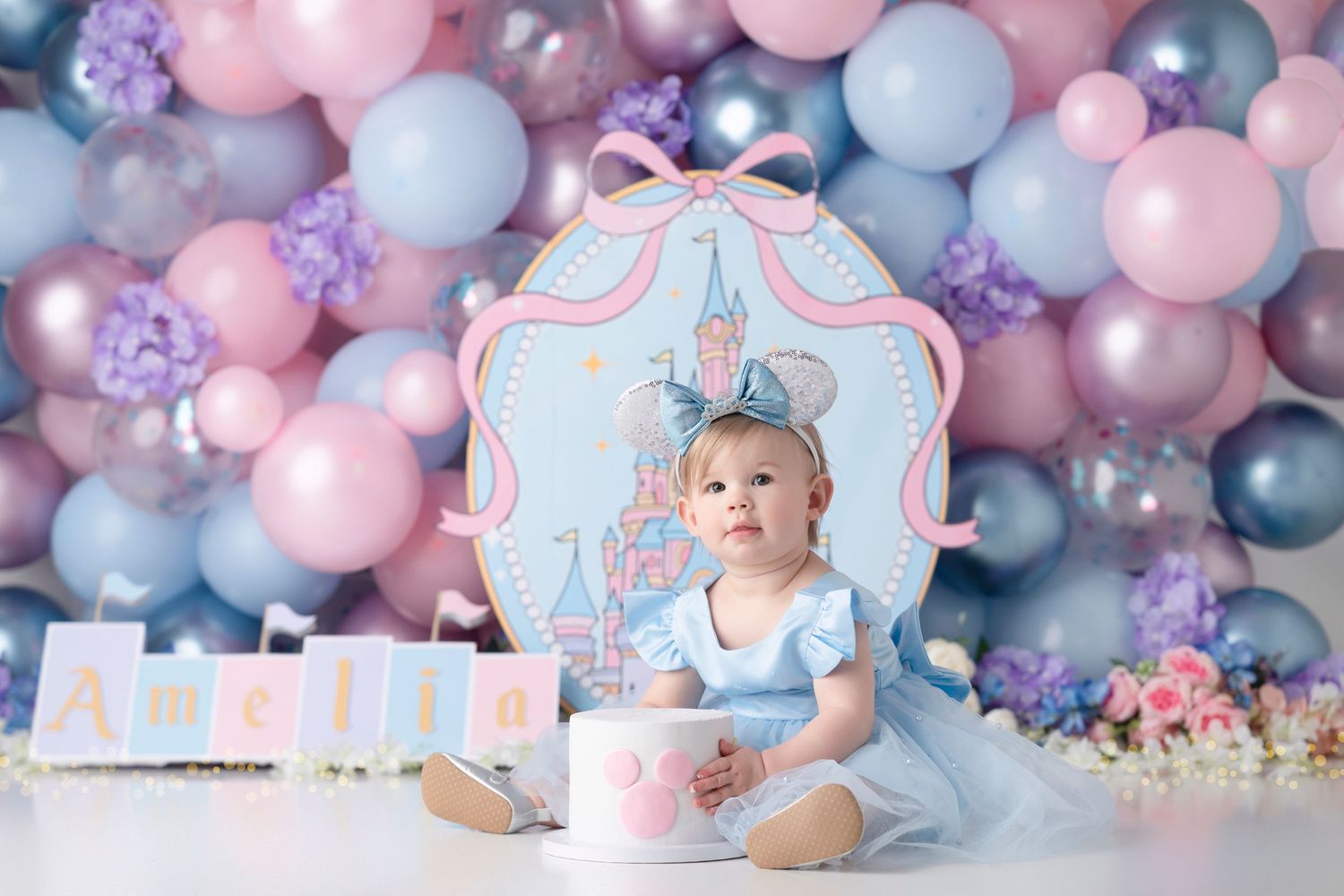 Baby girl in blue dress sits by castle backdrop with pink and blue balloons for a Disney-themed first birthday cake smash.