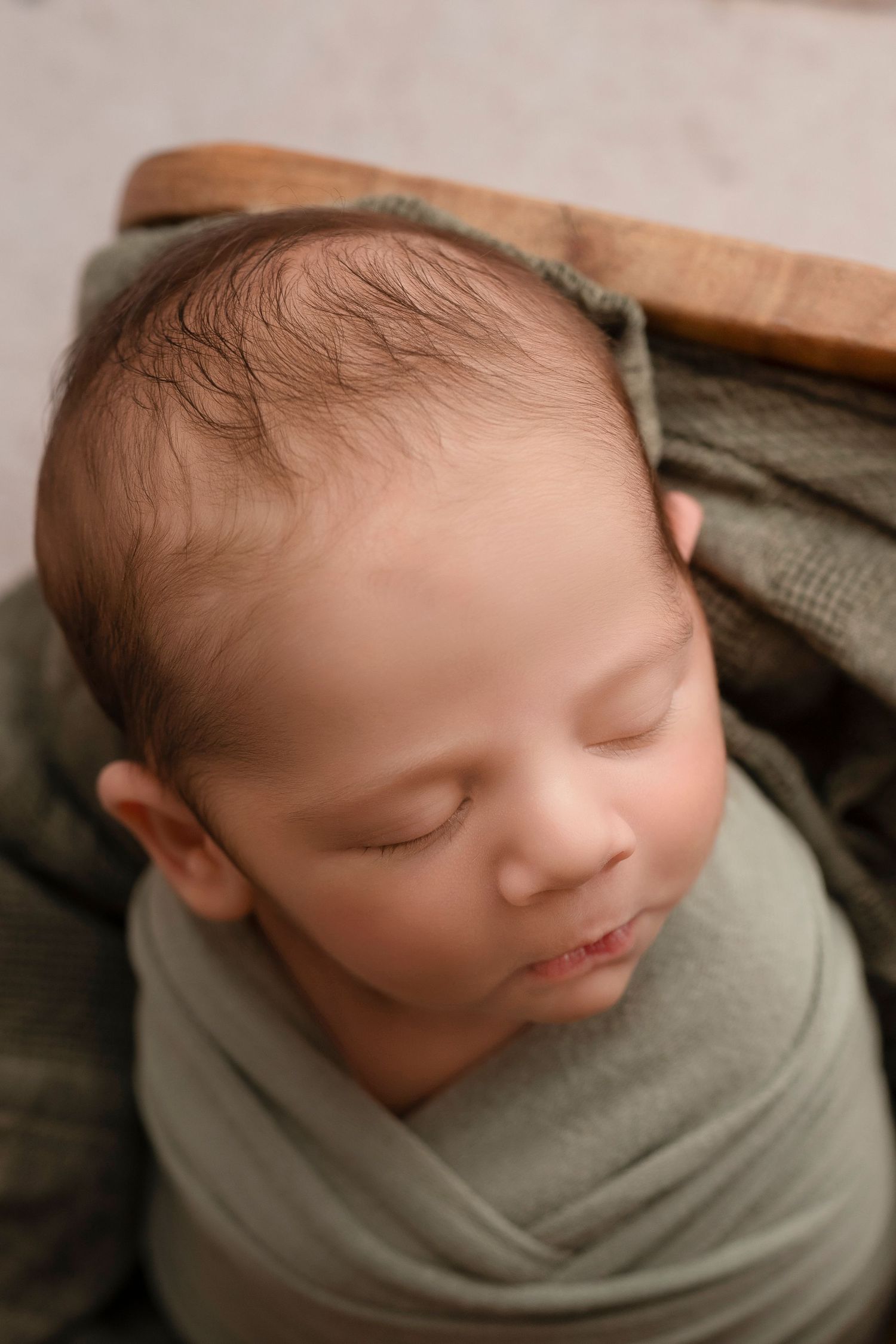 Sleeping newborn wrapped in sage green fabric lying on wooden surface in artistic portrait series.