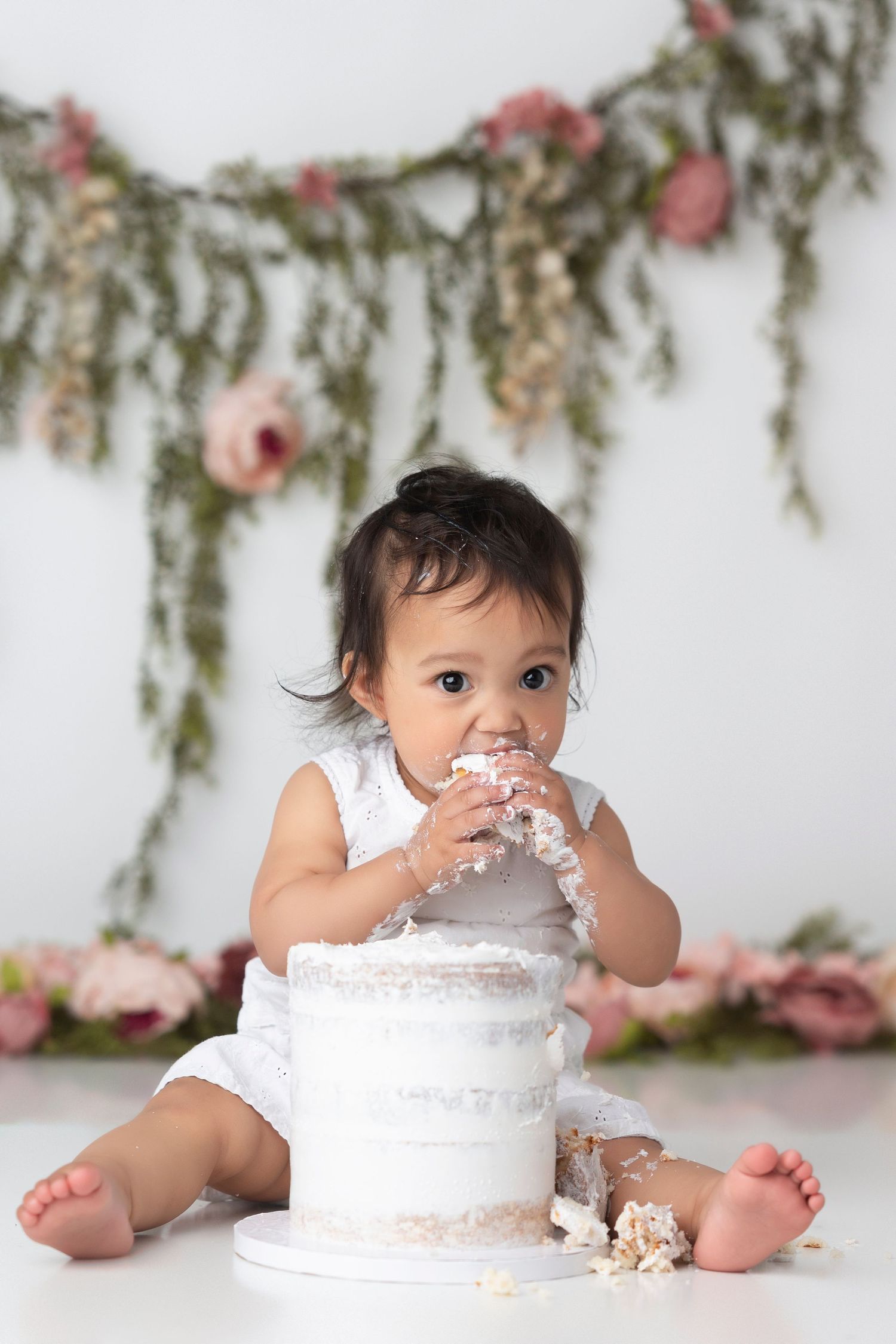 Baby in white dress smashes white birthday cake during cake smash photo session with floral backdrop.