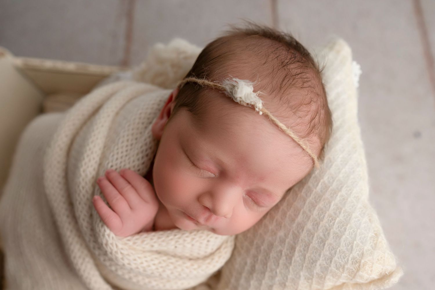 Newborn baby sleeps peacefully on white blanket wearing delicate headband and wrapped in soft swaddle.
