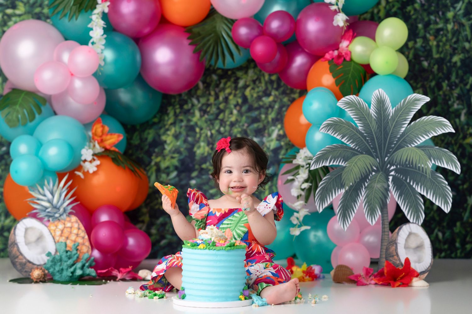 Colorful balloon garland with tropical palm leaf decorations and festive party setup against green foliage background.