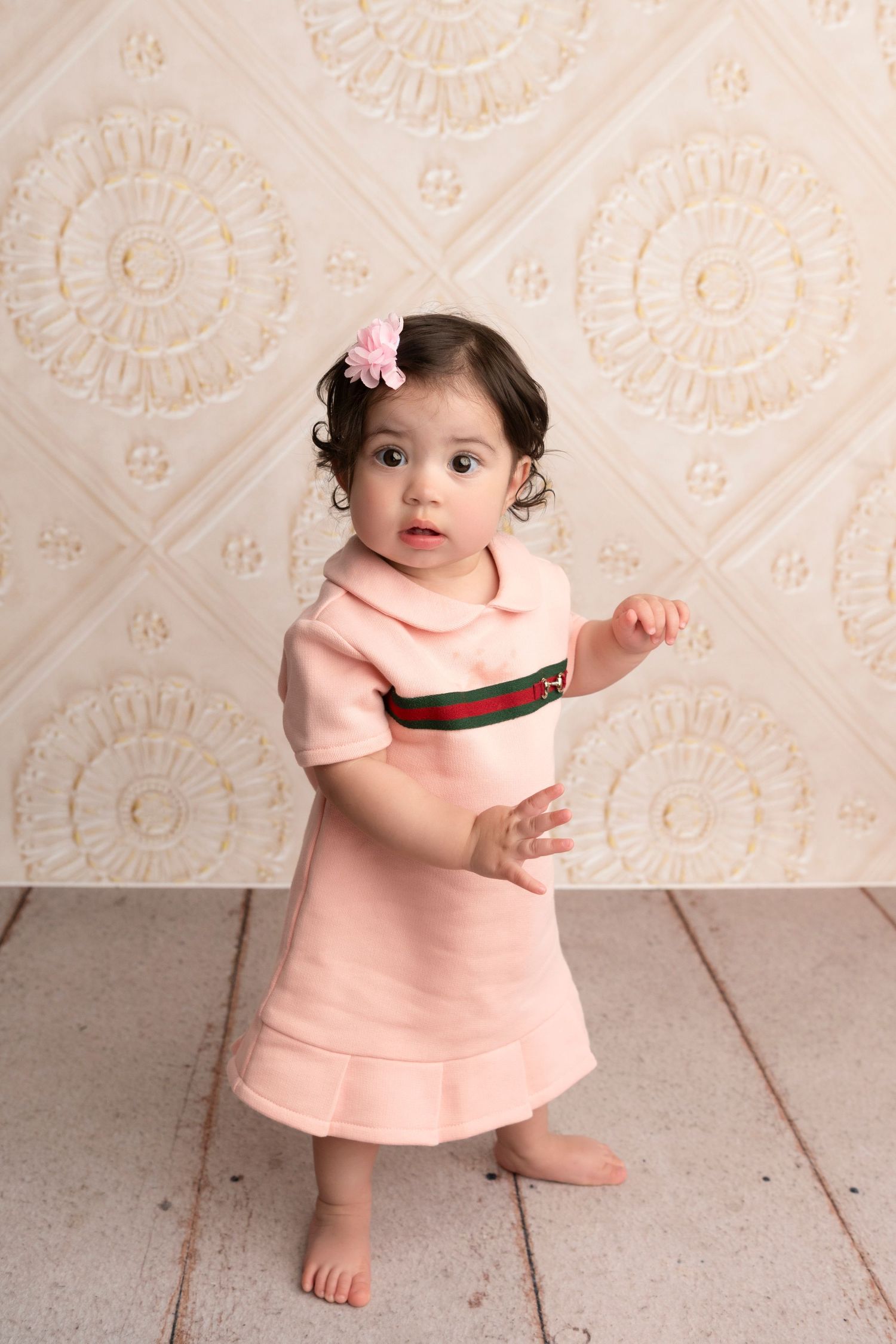 Sweet toddler in pink dress plays and poses against ornate cream-colored wall with floral pattern backdrop.