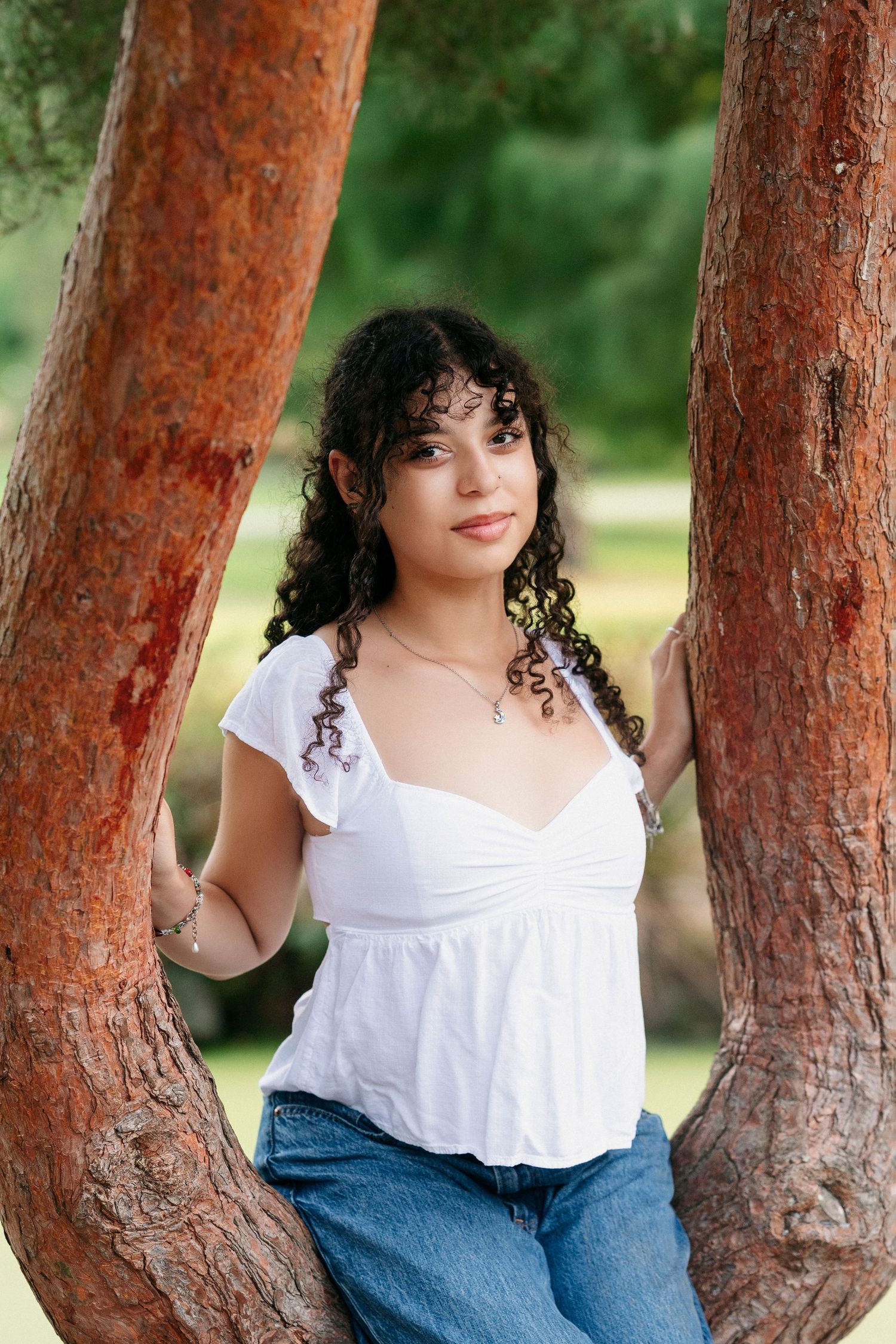 A portrait of someone with curly dark hair wearing a white top and jeans, sitting between two reddish tree trunks.