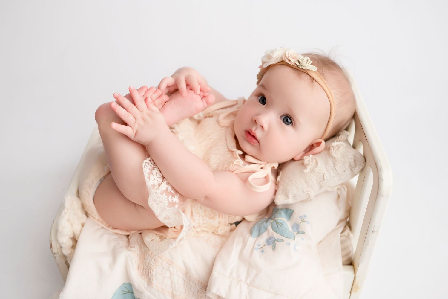 A sleeping baby wearing a cream-colored dress lies on a white blanket in a peaceful pose.