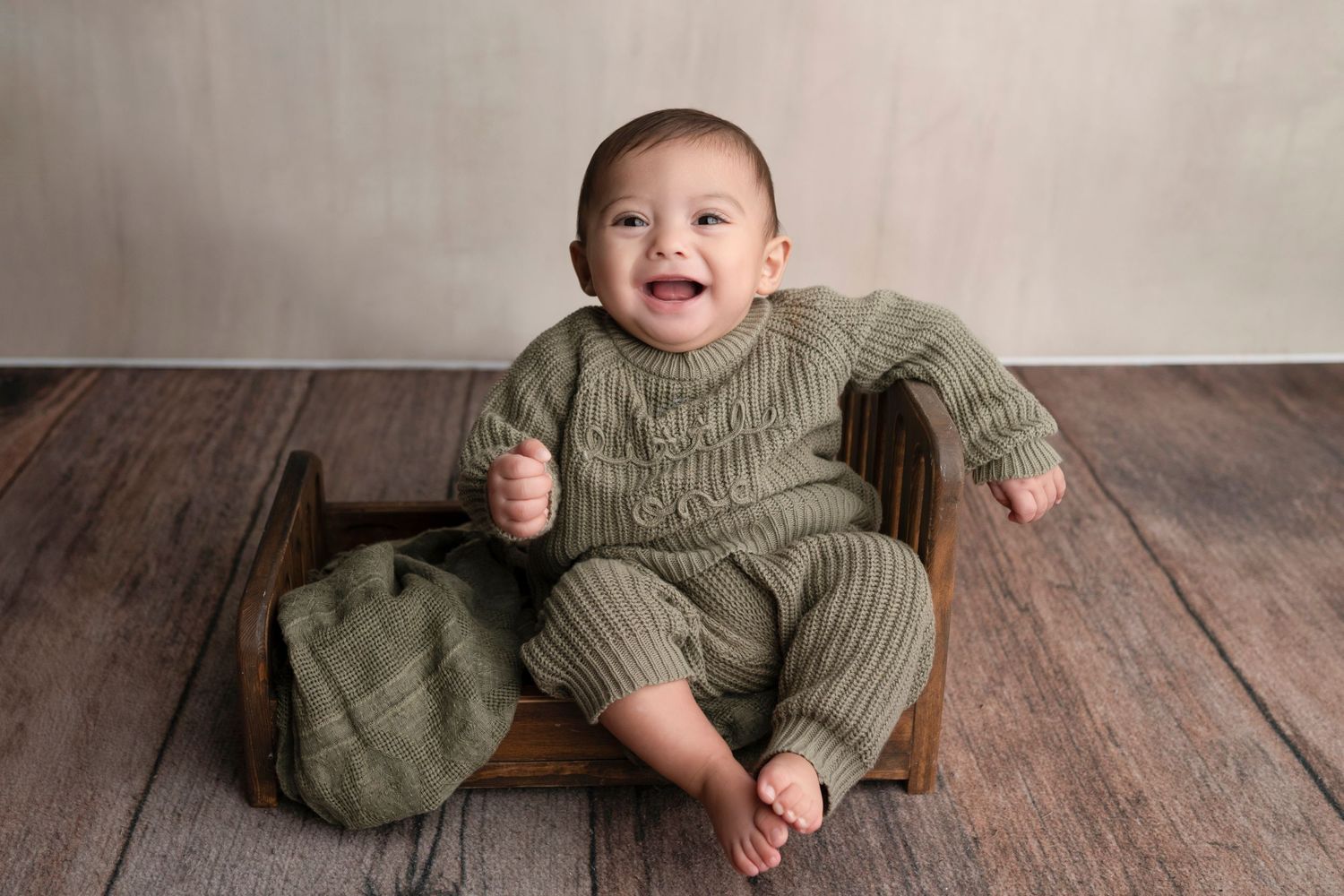 Smiling baby in green knit outfit sitting in a small wooden chair on a wood floor.