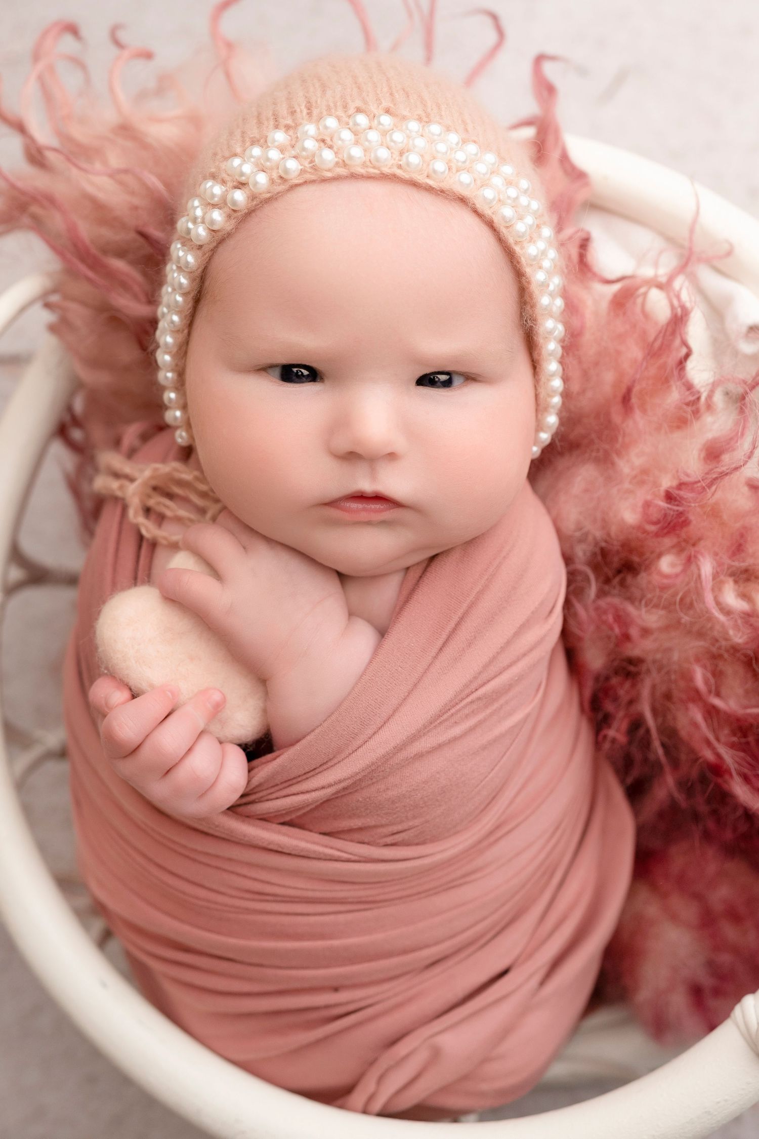 Newborn baby girl swaddled in pink wrap wearing pearl bonnet, posed in white basket with pink fluffy decor.