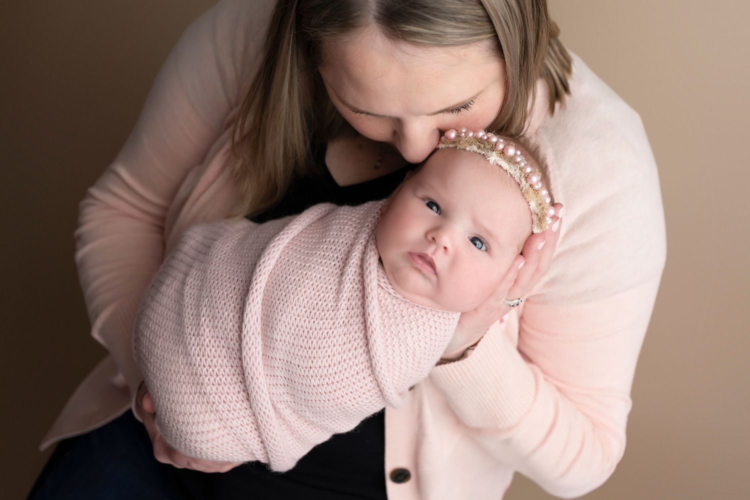 Mother holding swaddled newborn baby in pink wrap and pearl headband during professional studio portrait session.