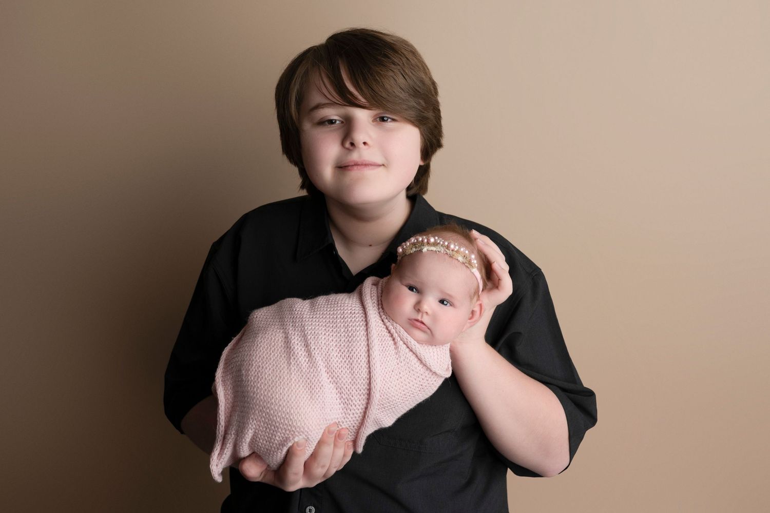 Older boy in black shirt proudly holds swaddled newborn baby girl wearing pink headband against beige backdrop.