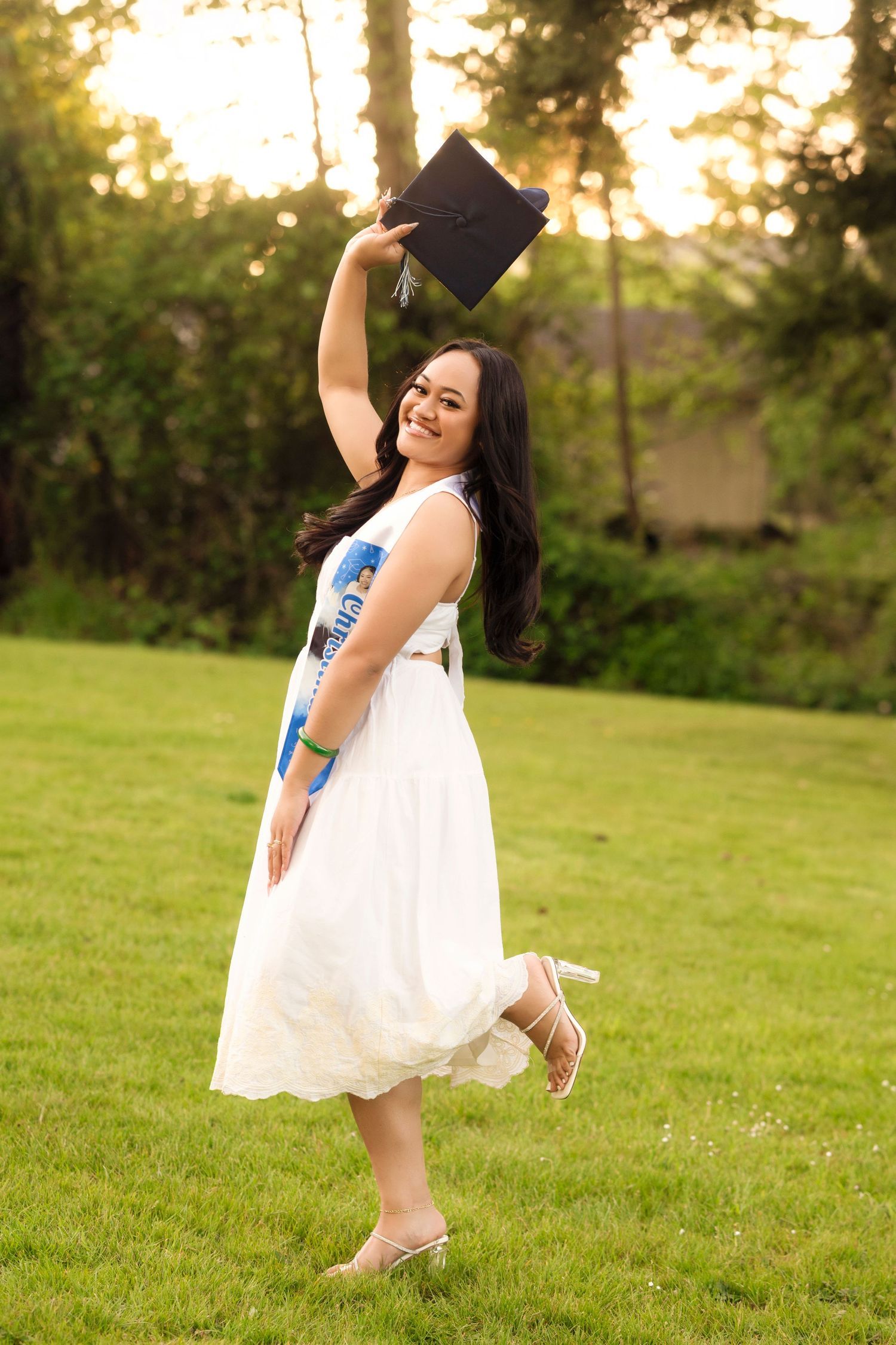 Graduate in white dress poses joyfully in outdoor setting during golden hour sunset.