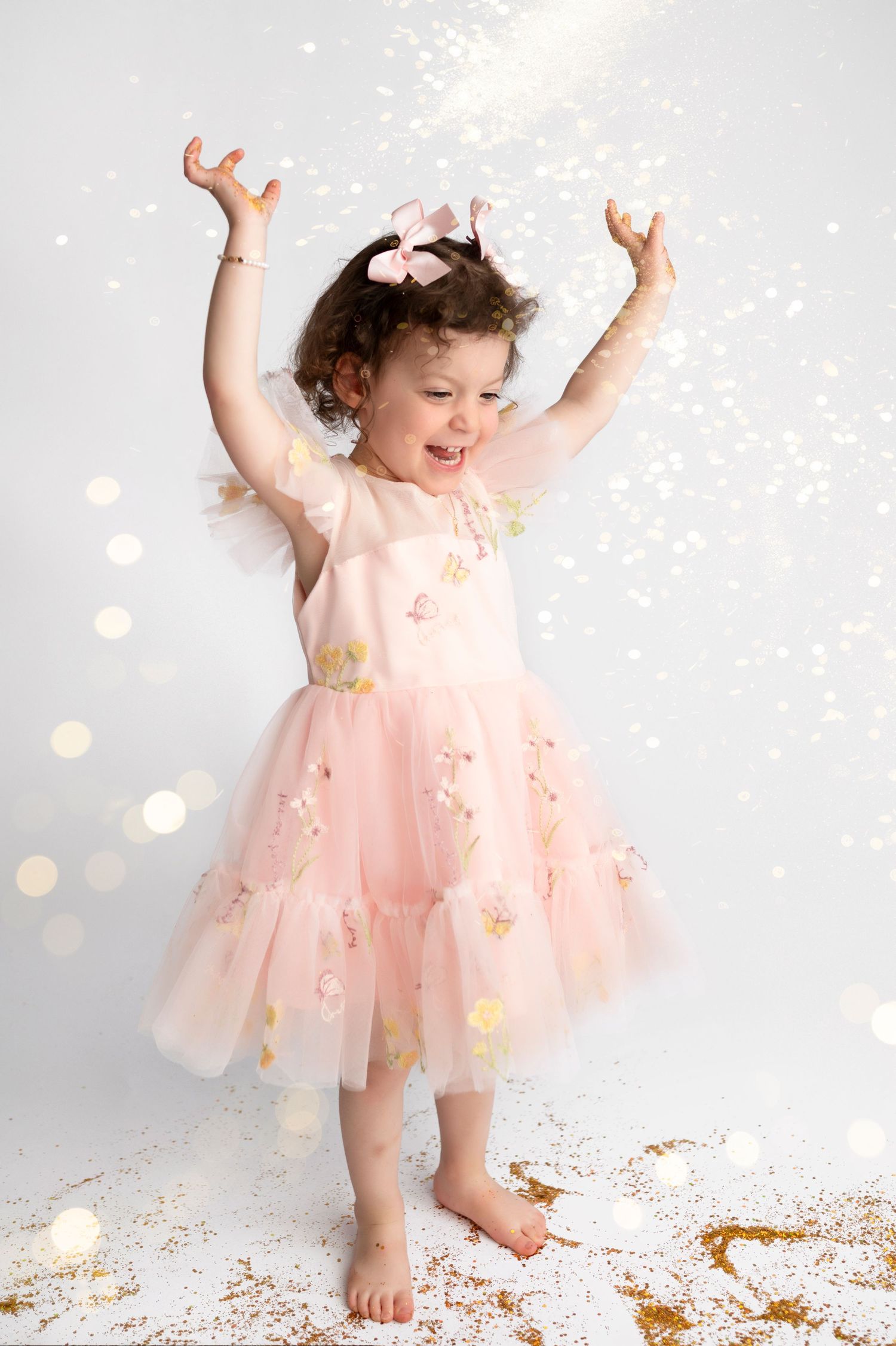 A small child in a pink tulle dress dances joyfully among scattered gold confetti against a light background.