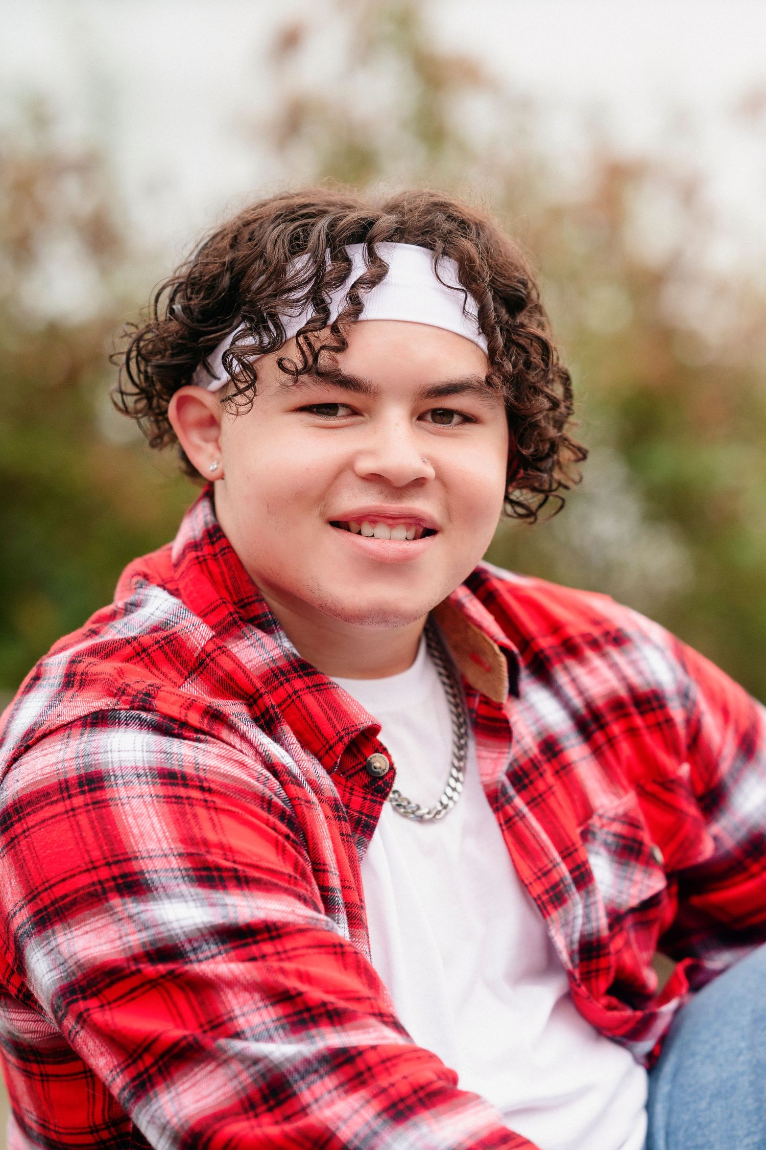 A casual outdoor portrait of someone wearing a red and white plaid flannel shirt with curly hair and a bright smile.