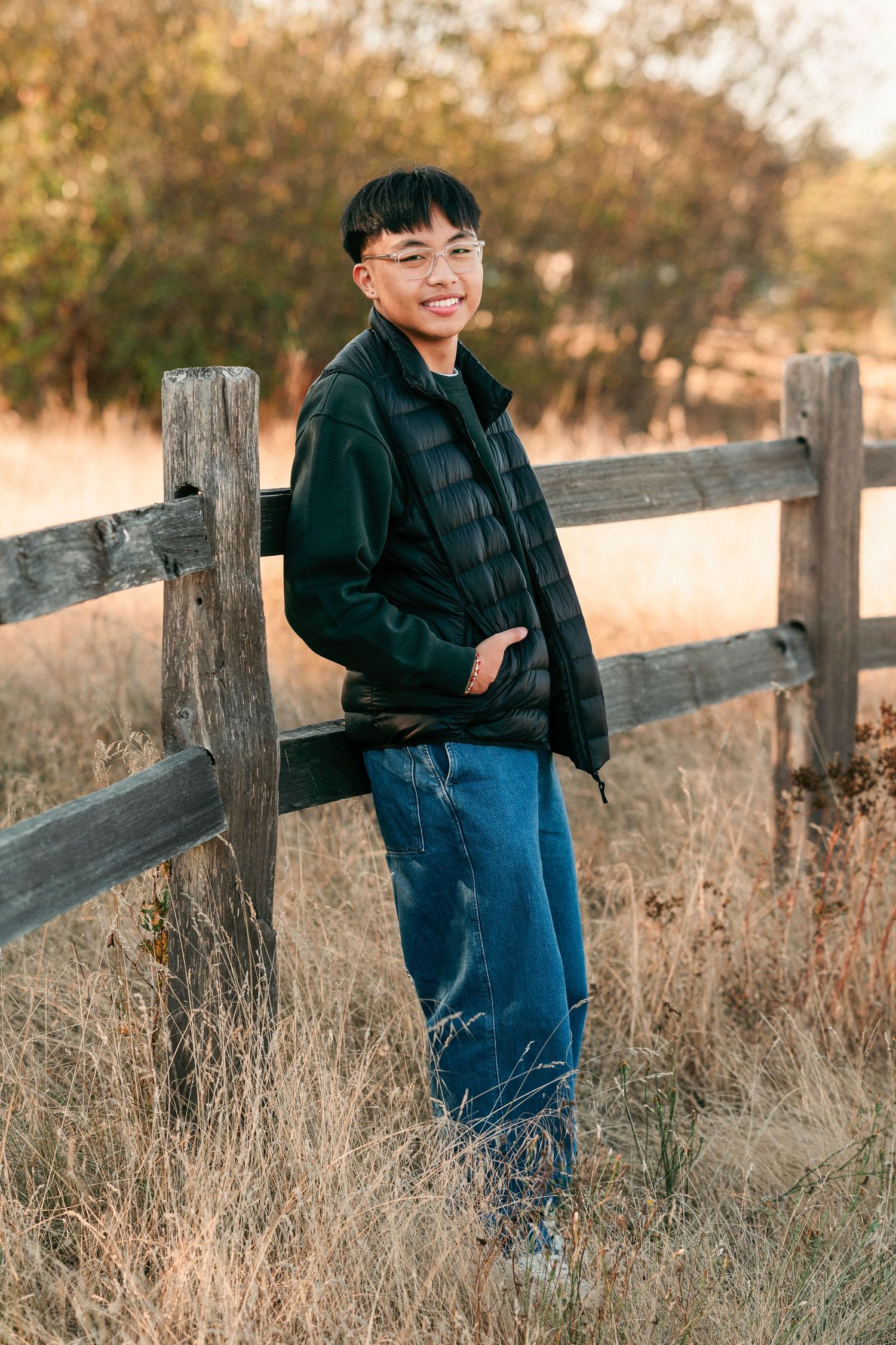 A figure in a dark jacket and jeans leans against a rustic wooden fence in a golden autumn field at sunset.