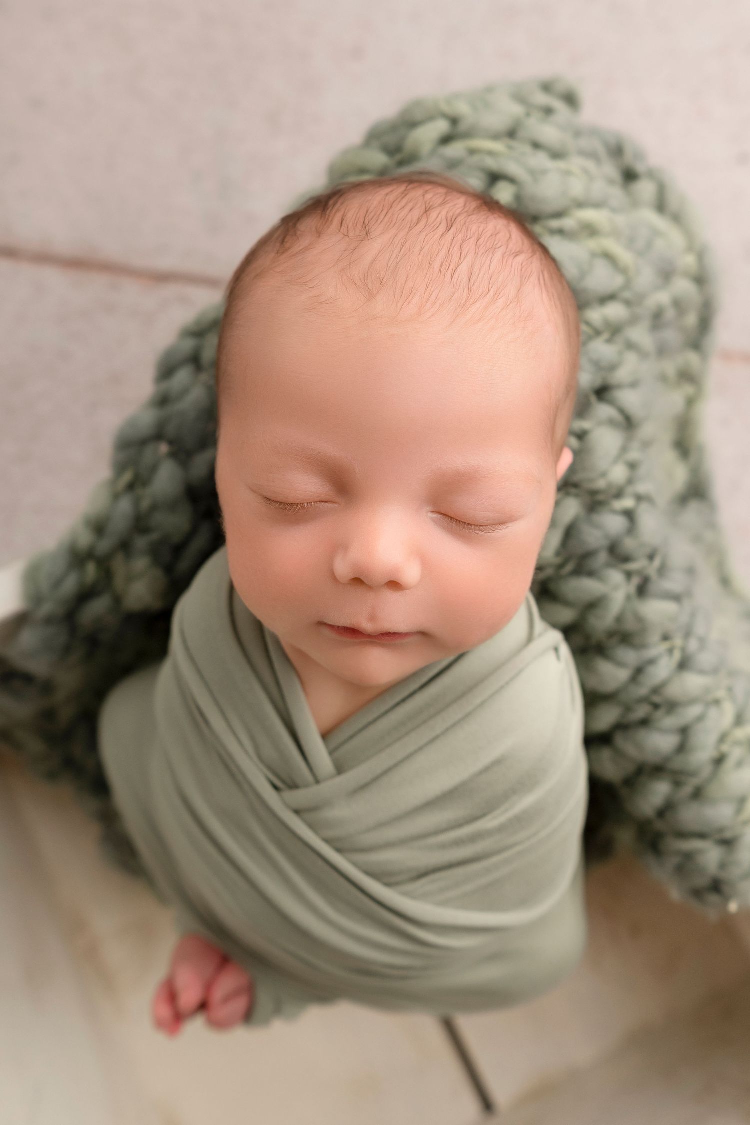 A sleeping newborn baby wrapped in sage green fabric against a soft textured background.