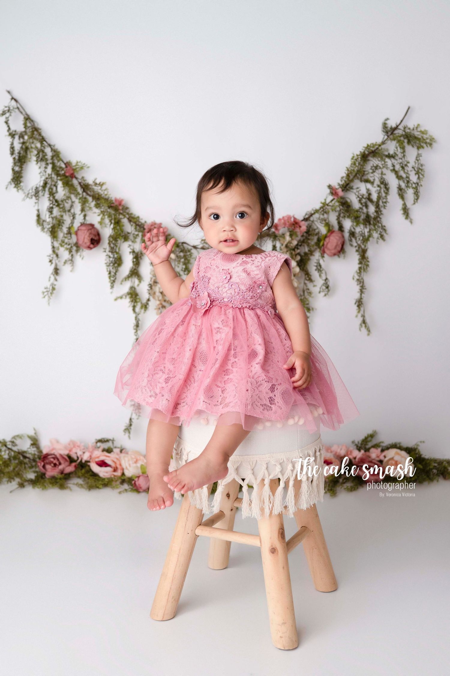 A baby in a pink lace dress sits on a wooden stool with evergreen branches decorating the background.