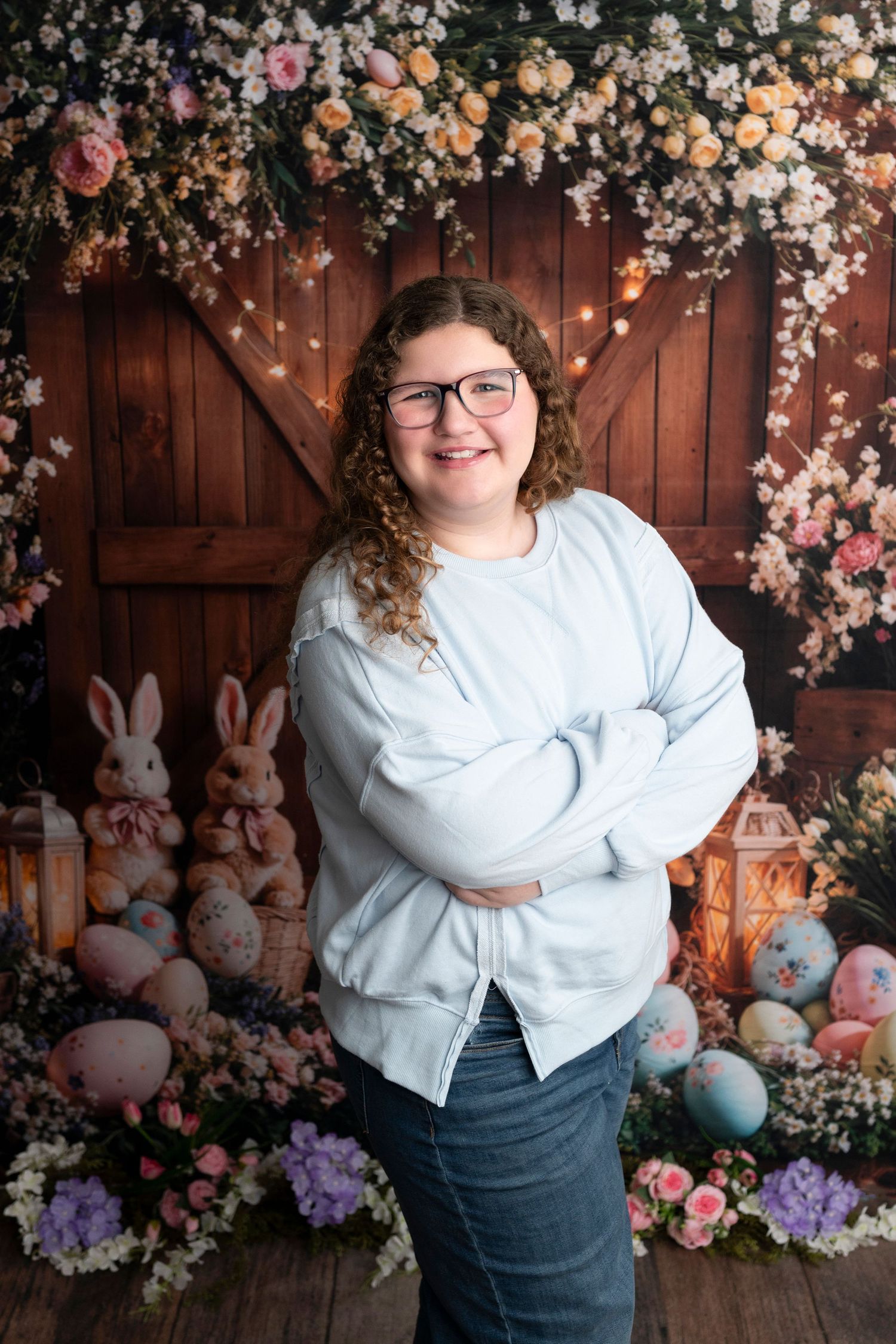 Young woman with curly hair and glasses smiles with arms crossed before a floral Easter backdrop with bunnies and eggs.