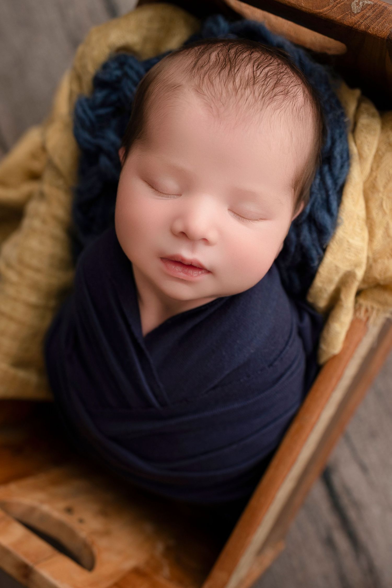 Sleeping newborn baby swaddled in navy blue wrap resting in a wooden box with mustard and blue knit blankets.