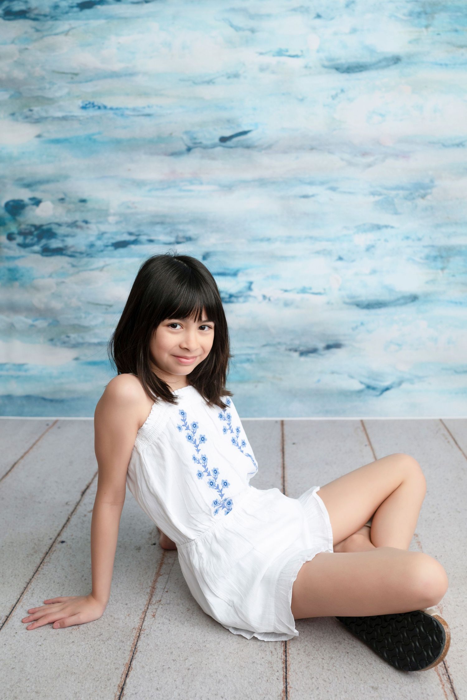 Young girl in white embroidered dress sitting on wooden floor against blue watercolor backdrop.