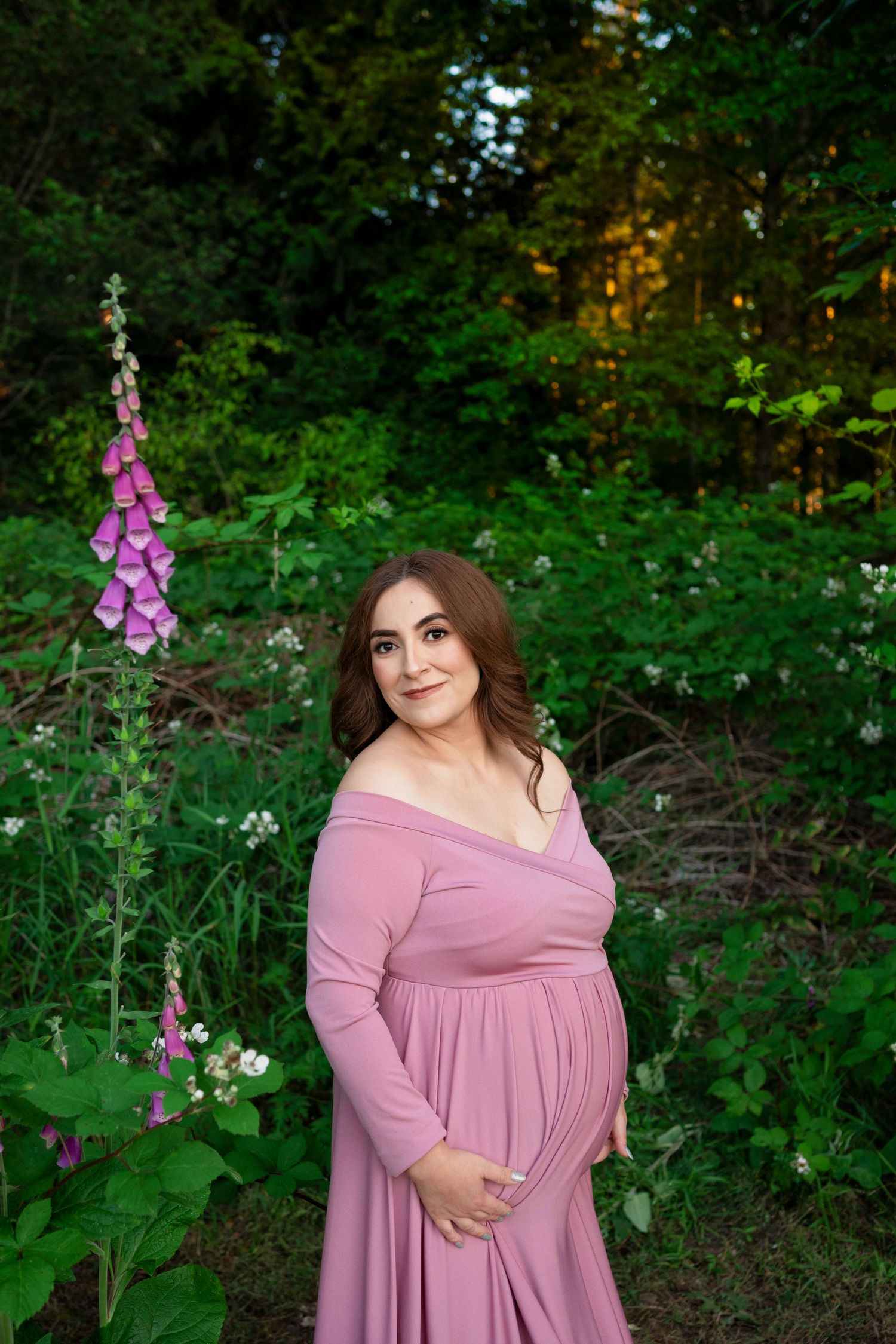 A person in a flowing pink dress poses gracefully outdoors among purple foxglove flowers and lush green foliage.