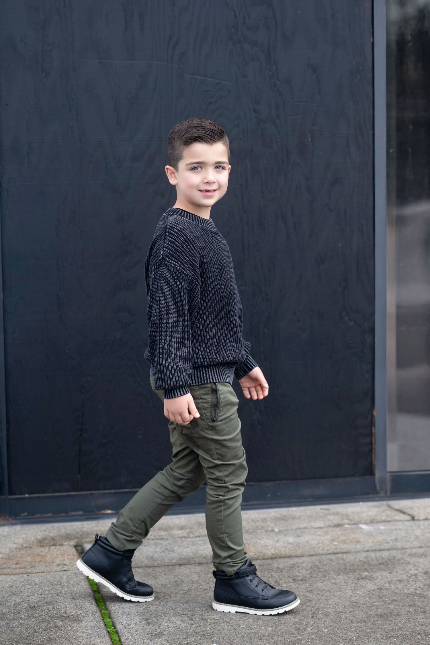 A person in a black sweater and olive green pants walks across a concrete surface wearing black sneakers.