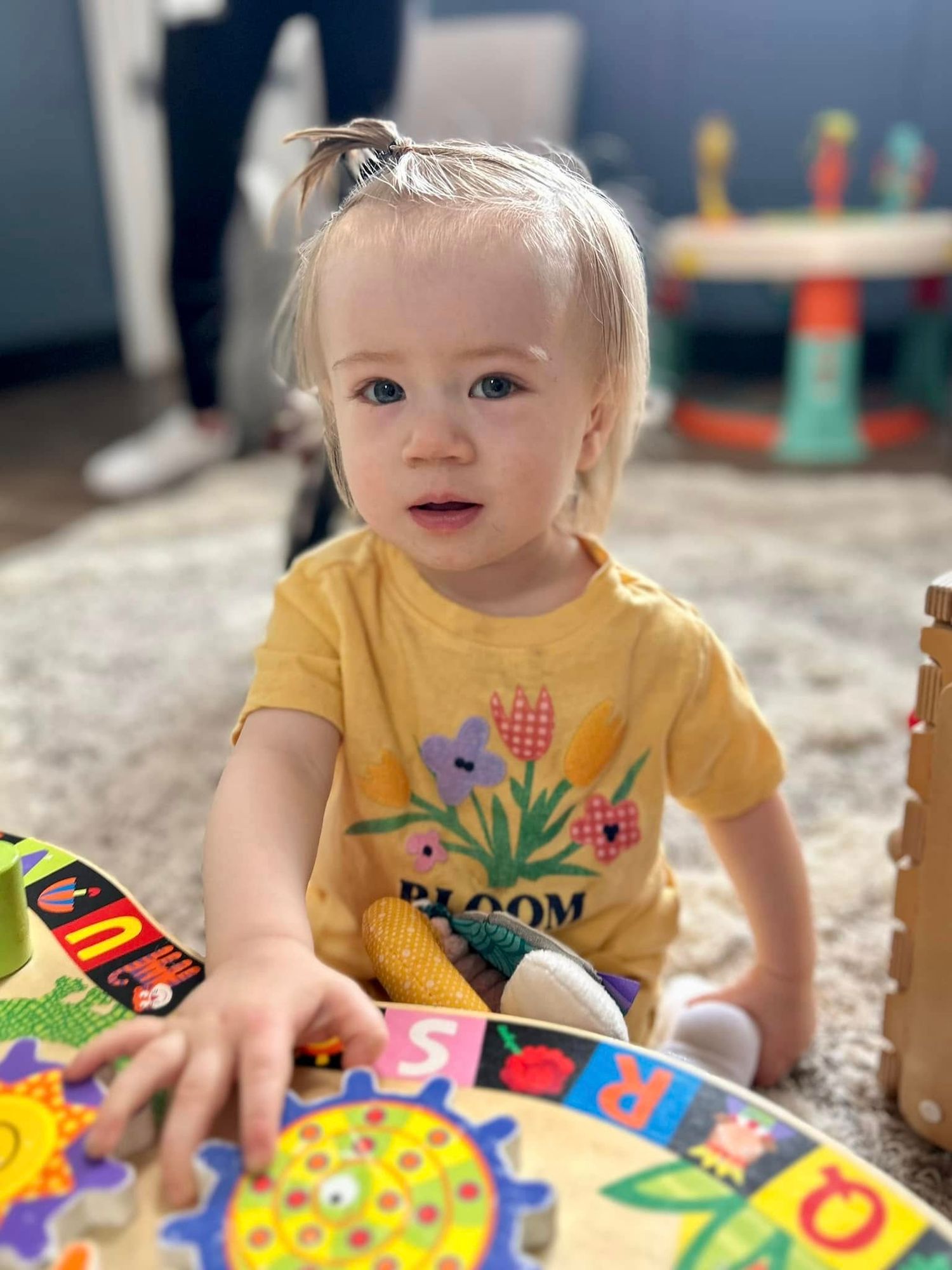Toddler in yellow Bloom shirt plays with colorful wooden toys on carpet in bright playroom.