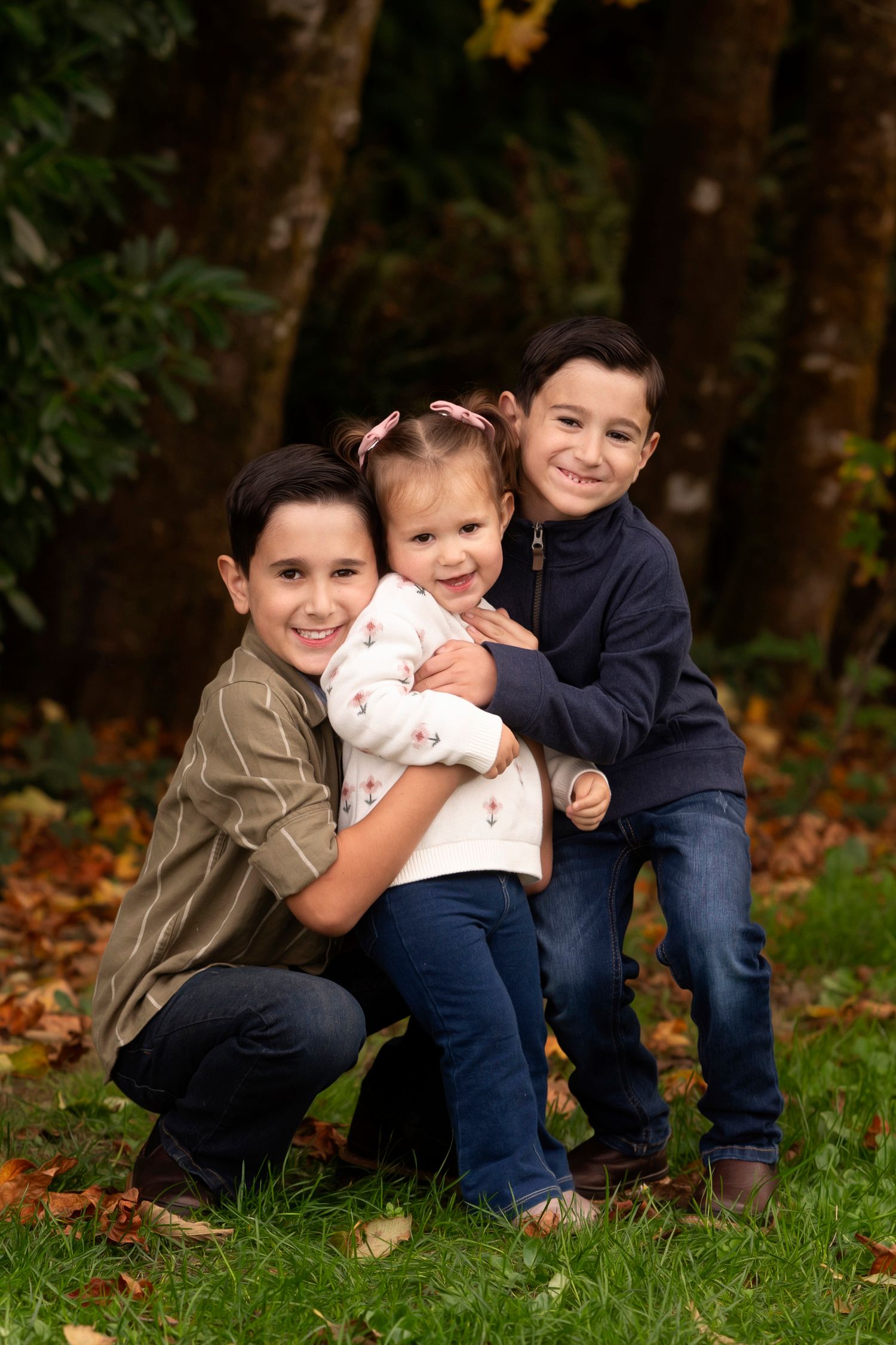Three children sit and play together on grass surrounded by fallen autumn leaves in a park setting.