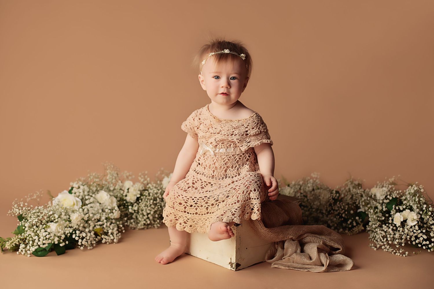 Baby in lace dress sits on wooden box surrounded by delicate floral arrangements against copper backdrop.