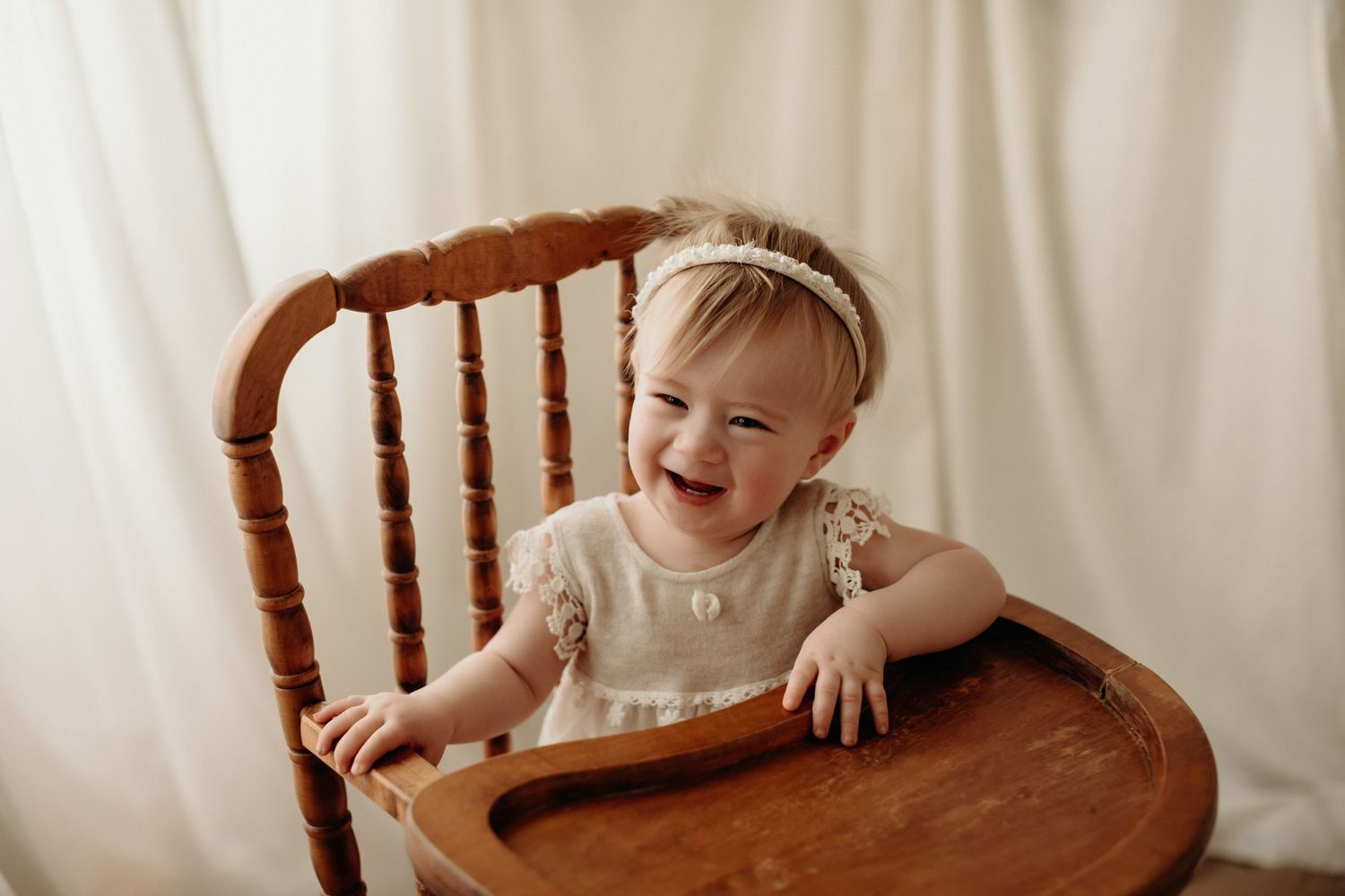 A joyful baby wearing a white dress sits in a wooden high chair against a light backdrop.