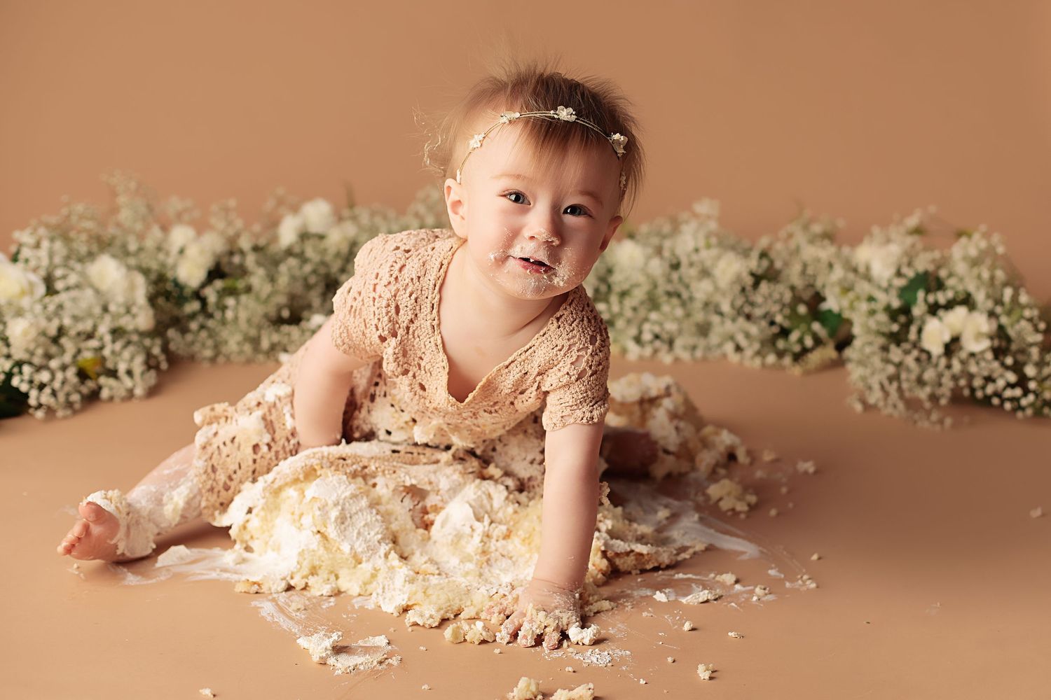 Baby in pink romper plays with flour and cake ingredients during messy cake smash photo session on brown backdrop.