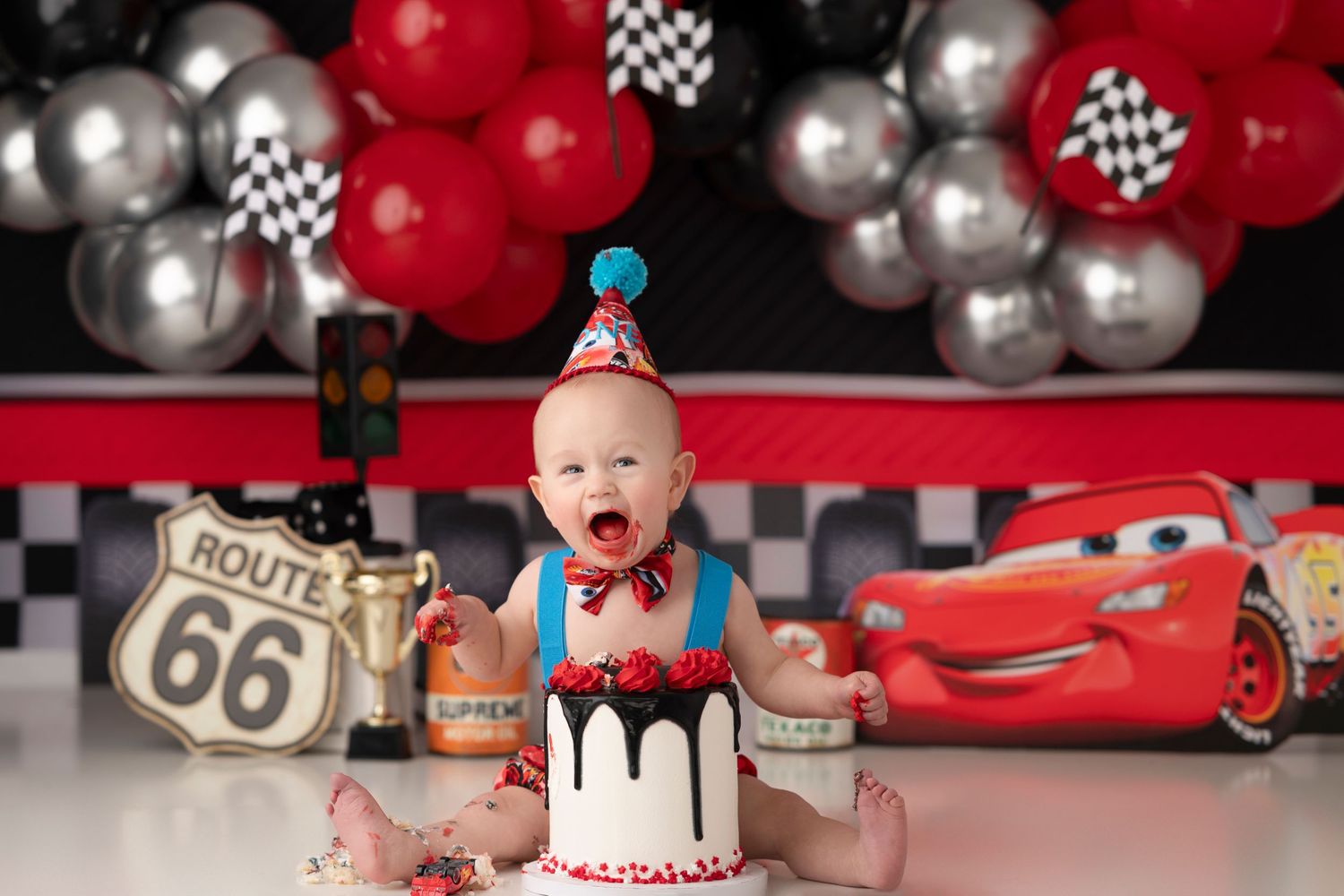 Baby's first birthday cake smash with Cars theme, red balloons, Route 66 sign, and Lightning McQueen backdrop.