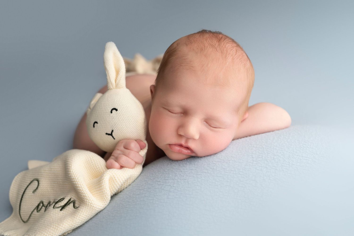 Newborn baby sleeping peacefully beside a cream knit bunny toy on a soft blue background.