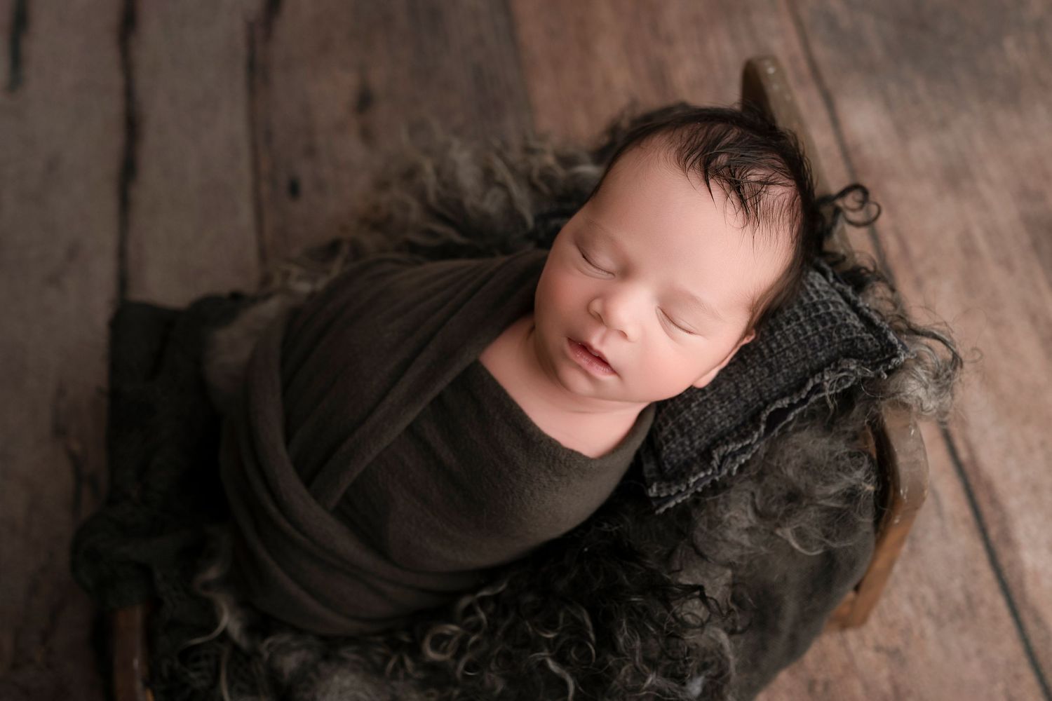 Peacefully sleeping newborn baby swaddled in brown wrap, nestled in a rustic wooden bowl with soft textured layers.