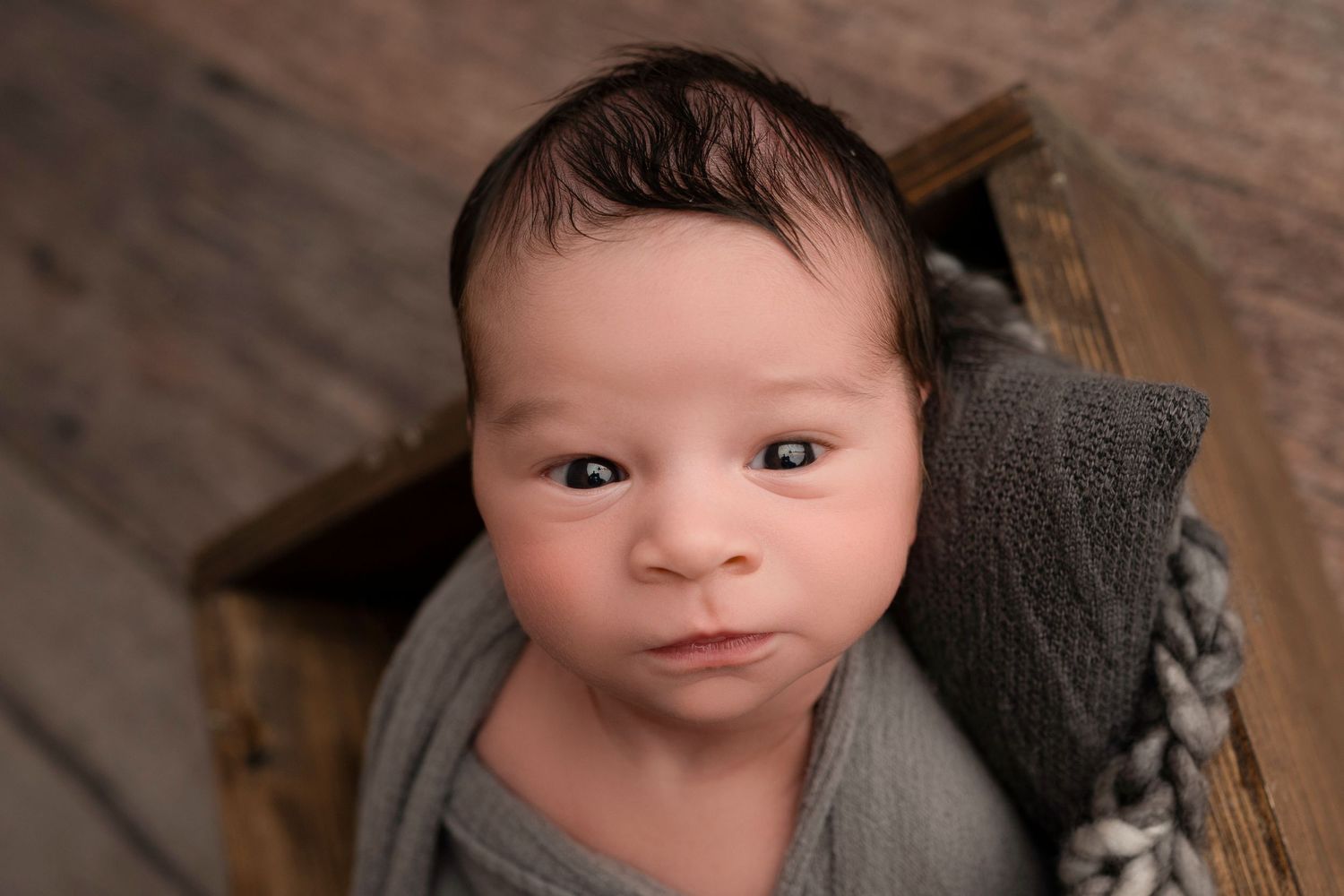Newborn baby wrapped in gray swaddle cloth, lying in a rustic wooden crate, looking up with alert dark eyes.