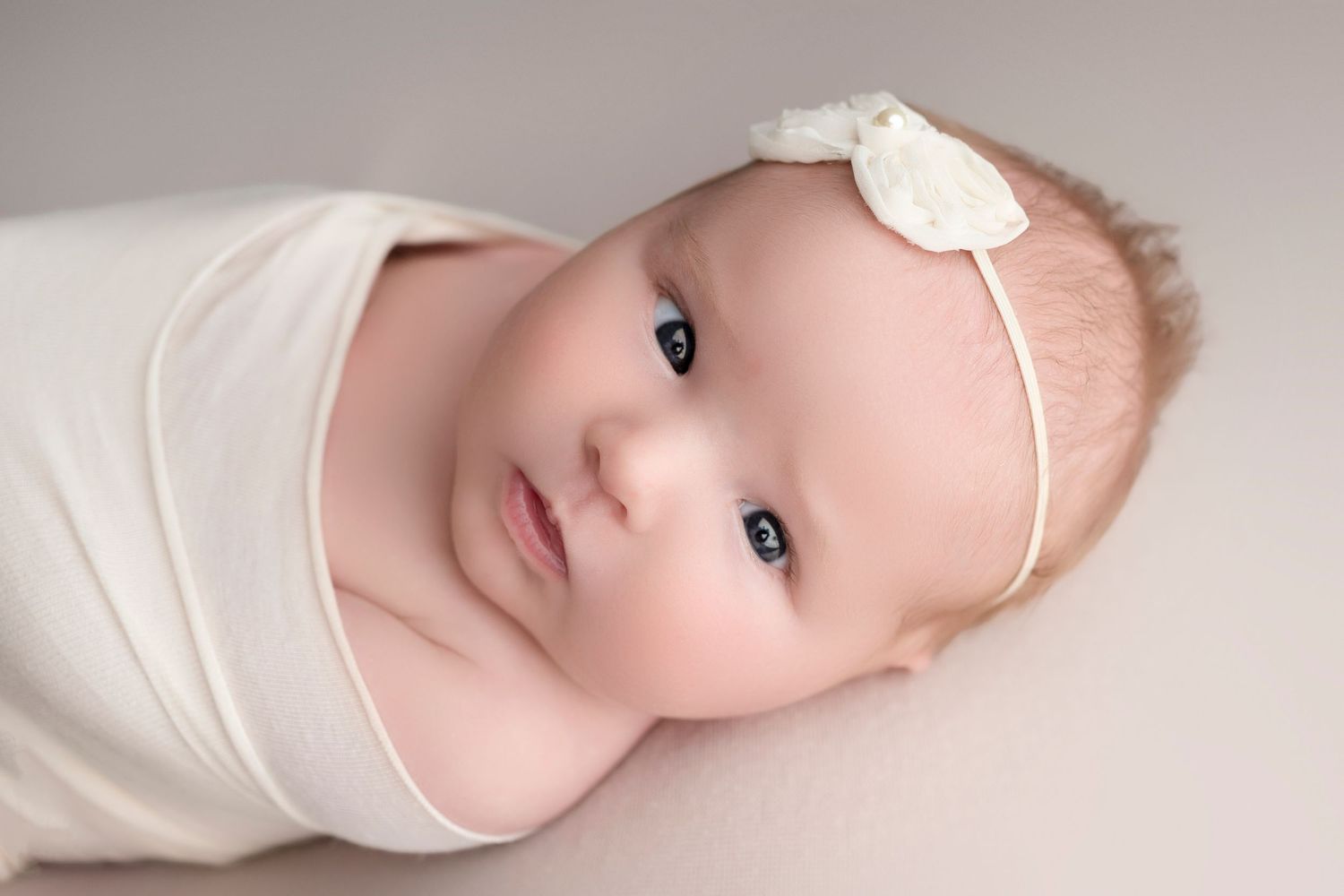Newborn baby swaddled in white wrap with floral headband, gazing softly at camera in studio portrait.