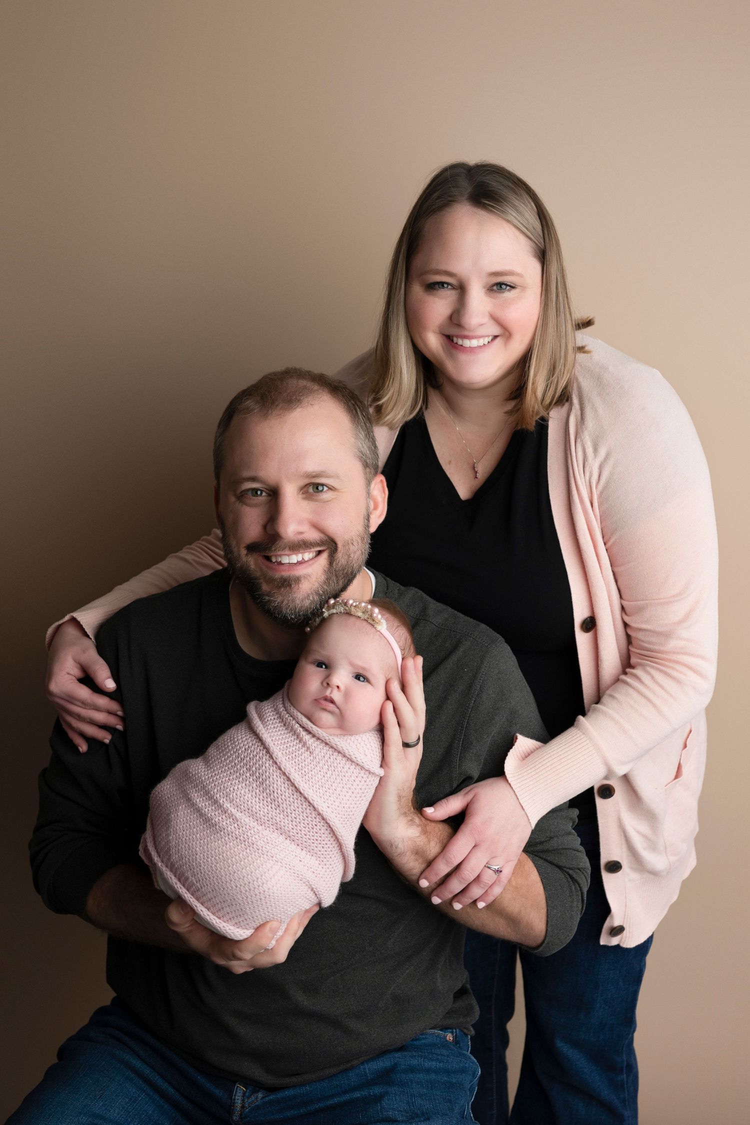 A smiling couple poses with their newborn baby girl wrapped in a pink swaddle blanket in a studio portrait.