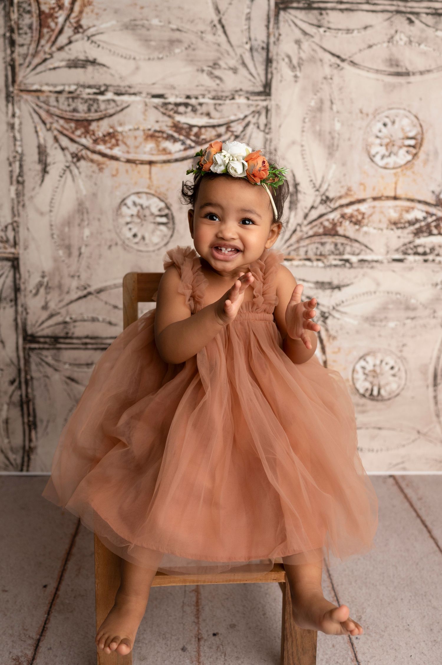 Smiling baby girl in a dusty rose tulle dress and floral crown, clapping hands while sitting on a wooden chair.