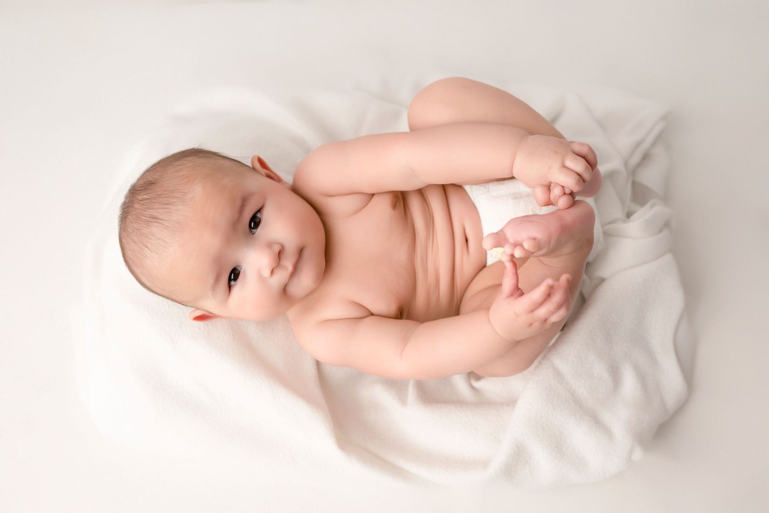 Adorable baby in a diaper lying on a white blanket, smiling and holding their feet up.
