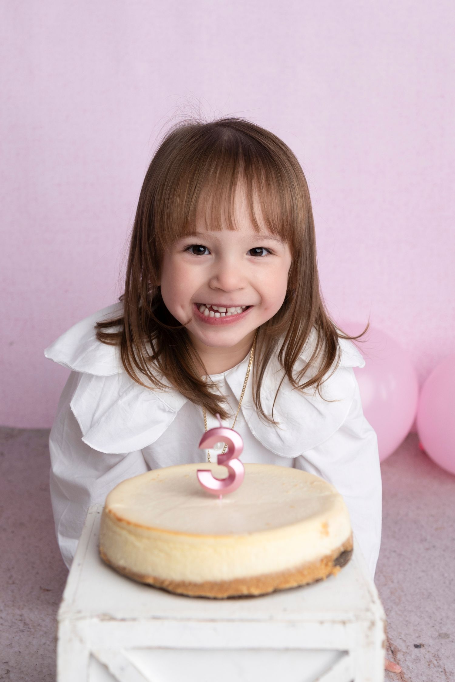 Smiling girl celebrating 3rd birthday with cheesecake and candle, pink balloons in background.