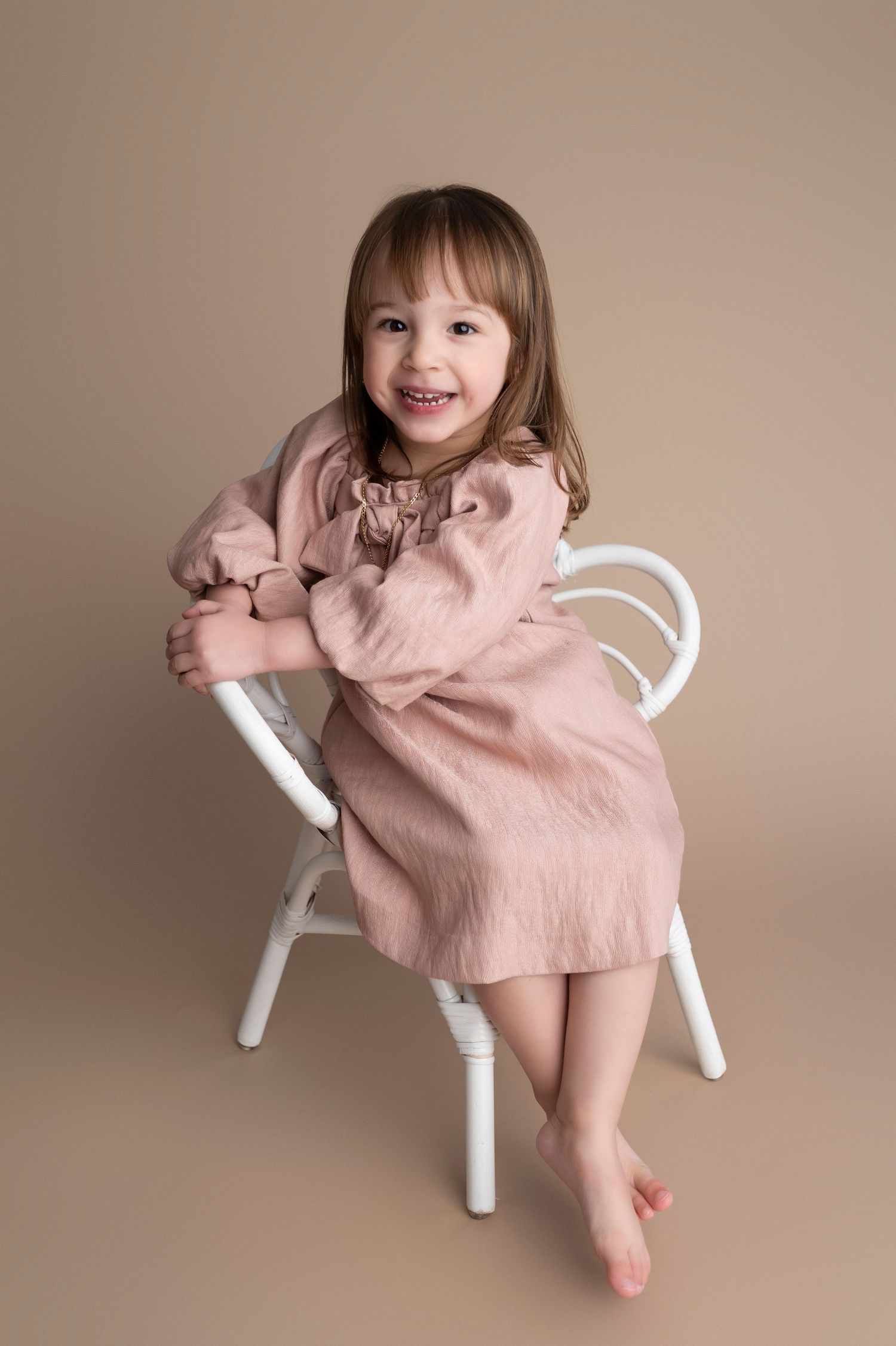 A smiling young girl in a pink dress sits barefoot on a white chair against a beige background.