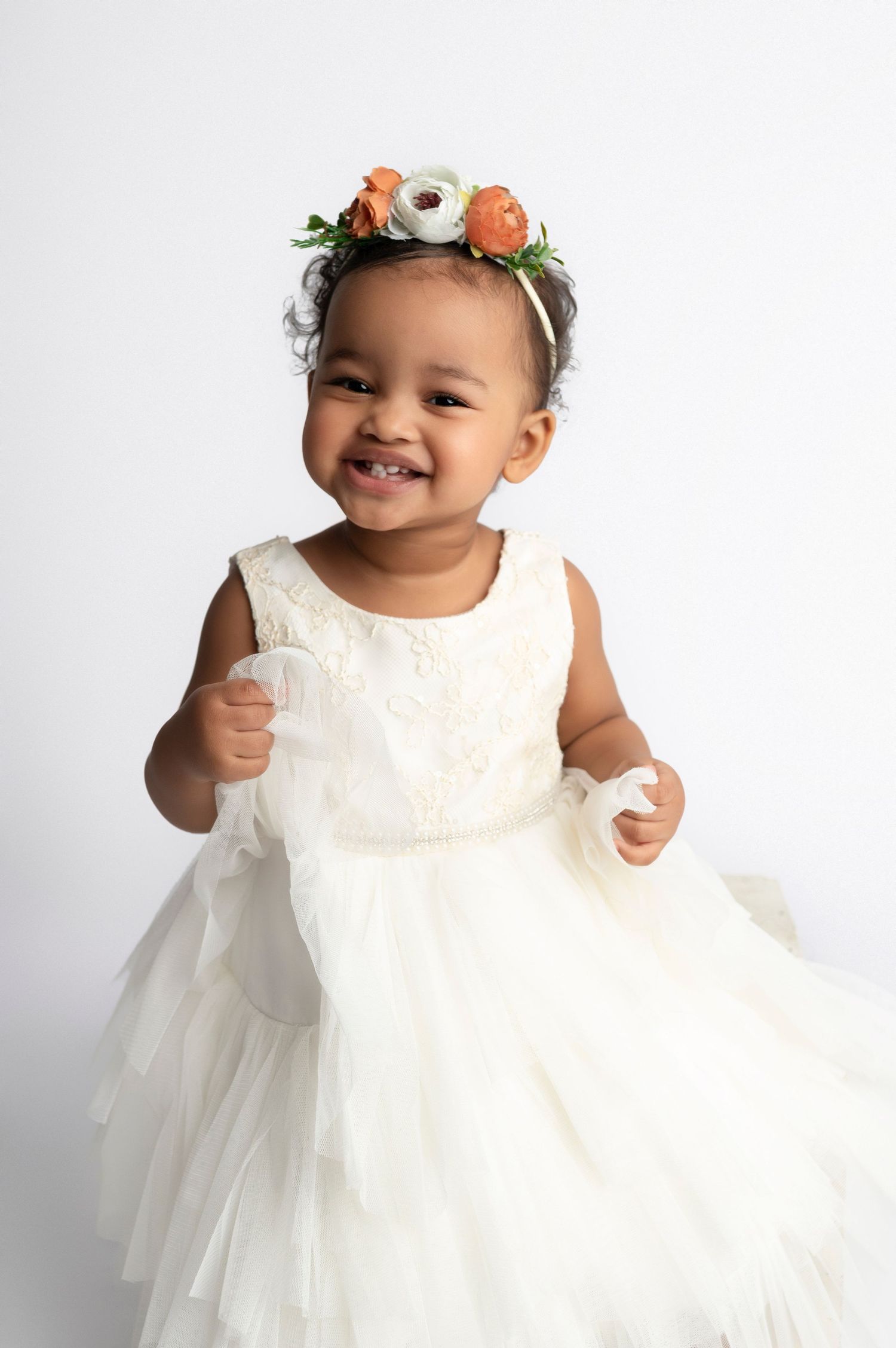 Smiling baby girl in white tulle dress and orange floral crown poses against white background.