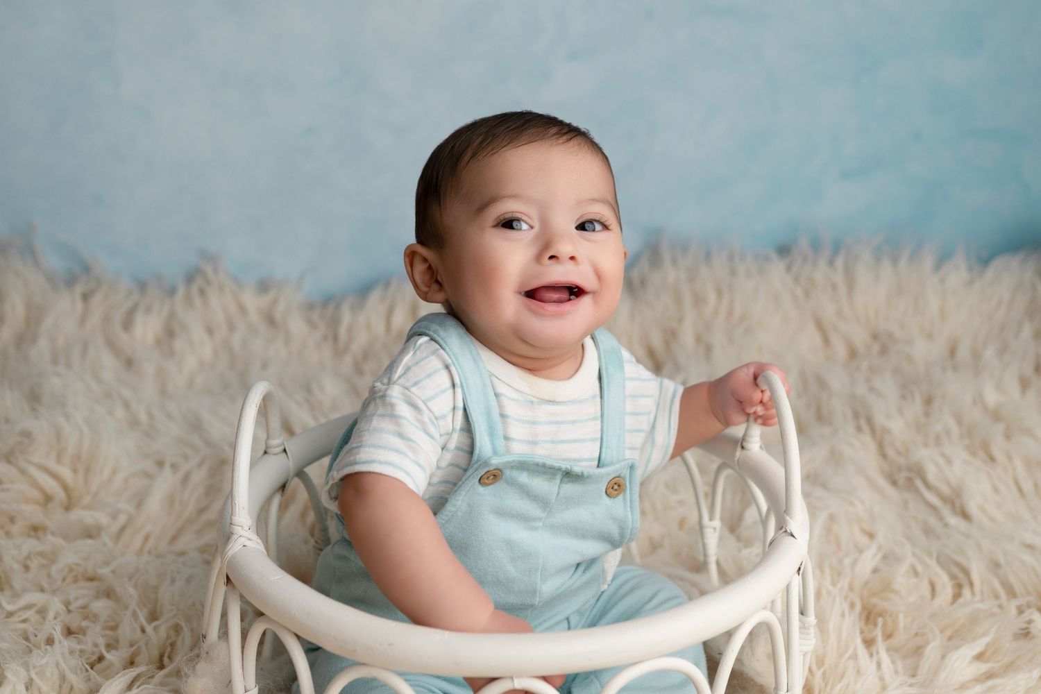 Smiling baby boy in blue overalls sitting in a white mini crib on a fluffy rug with a blue backdrop.