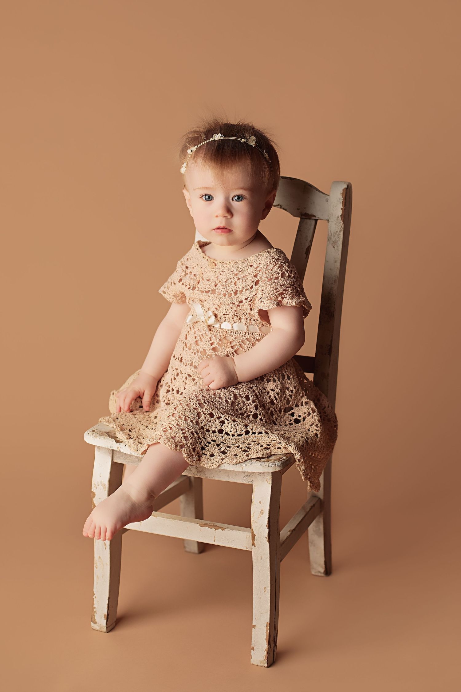 Small child in peach lace dress sits in antique white wooden chair against neutral brown backdrop.