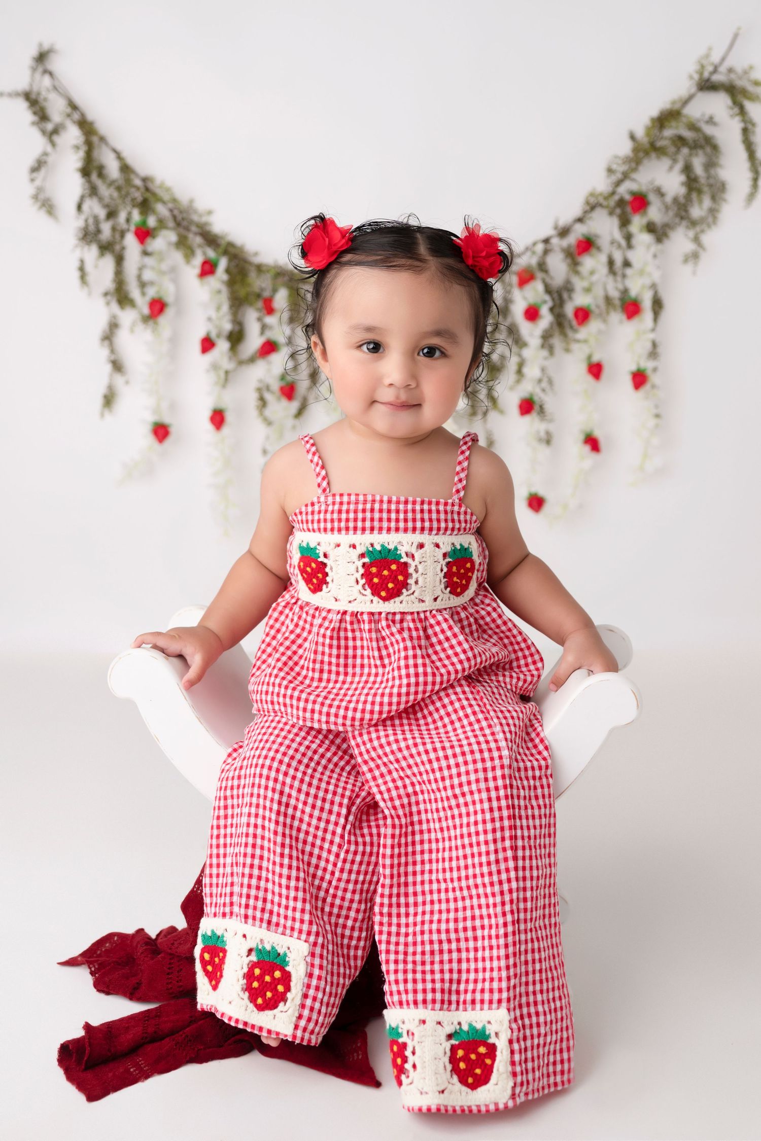 Toddler girl in red gingham strawberry smocked jumpsuit sits on white chair with floral strawberry backdrop.