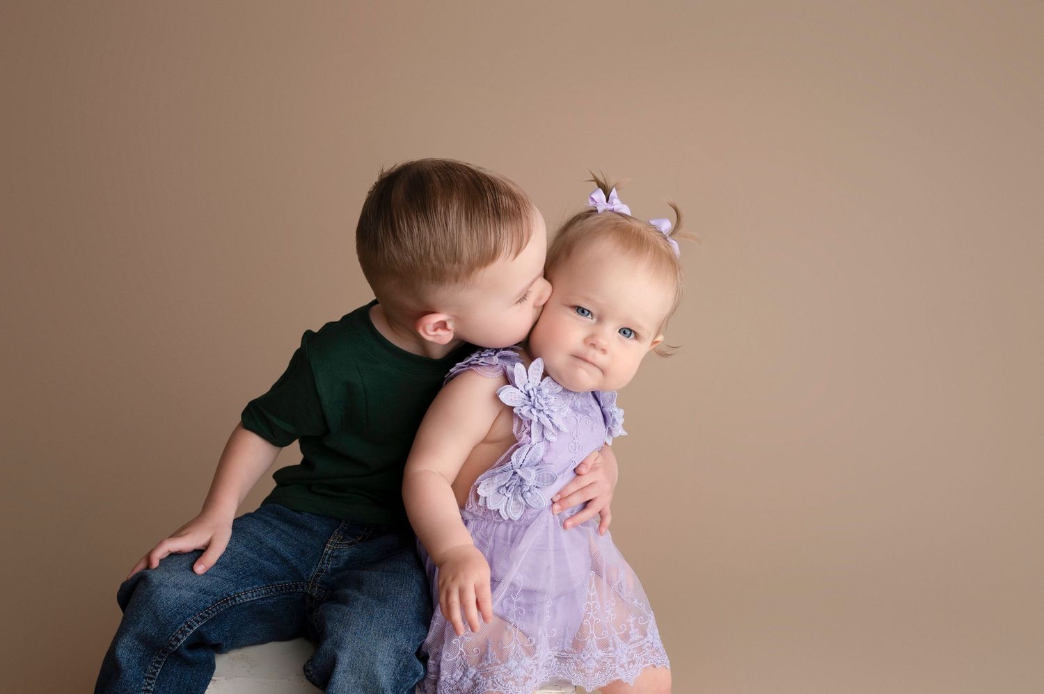 Sweet toddler boy in green shirt giving a gentle kiss to baby girl in purple lace dress on beige backdrop.