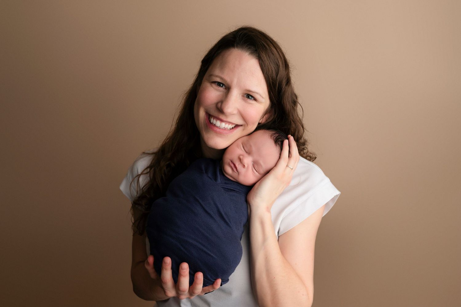 Smiling mother holding sleeping newborn wrapped in navy blue swaddle against a warm beige background.