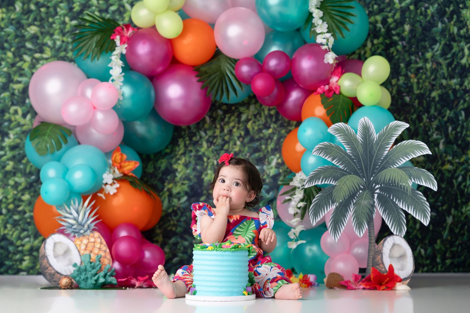 Colorful balloon garland with tropical decorations adorns a festive party setup against a green hedge wall.