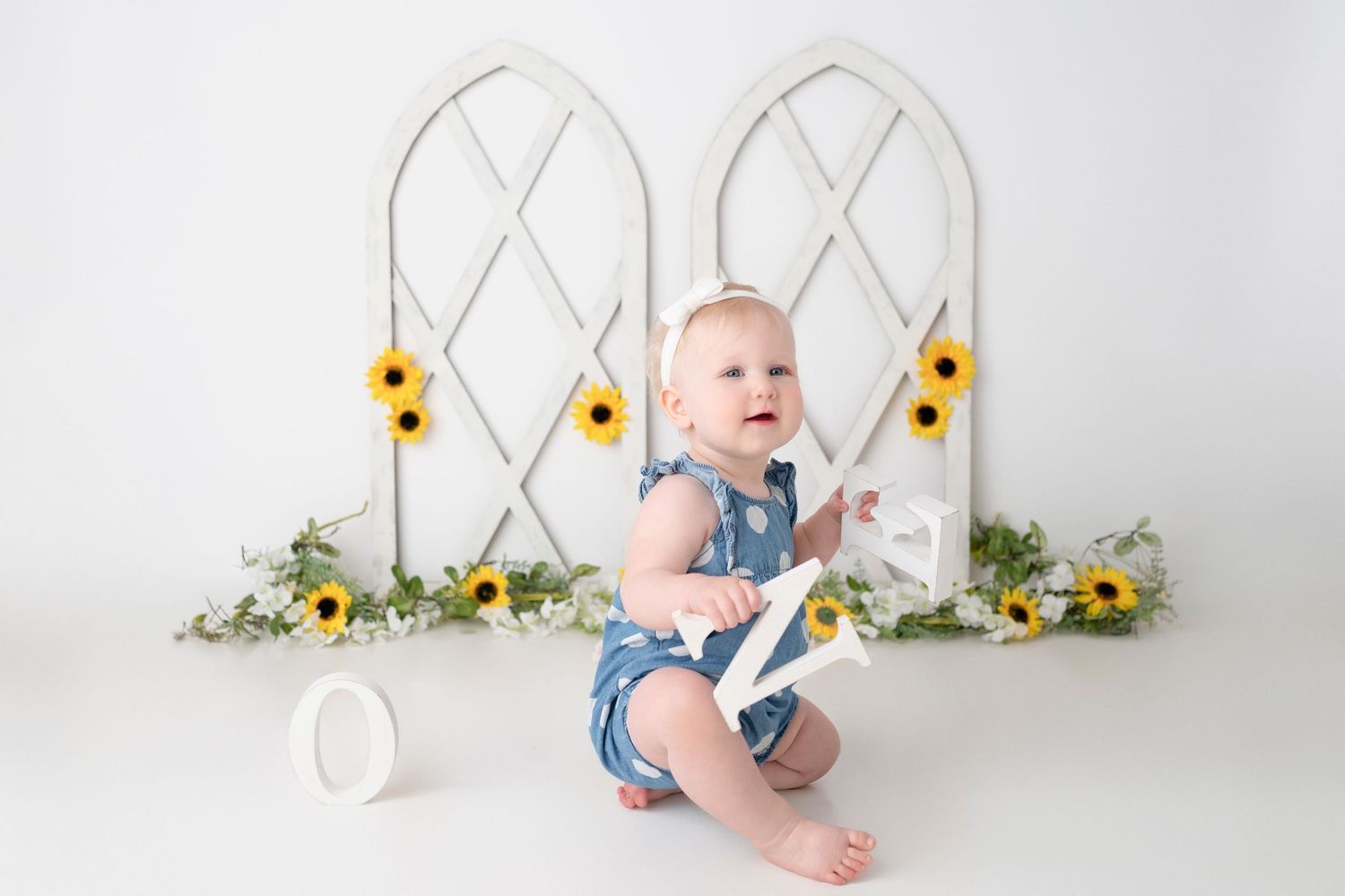 Baby in denim romper sits and plays against white lattice backdrop with yellow sunflowers.