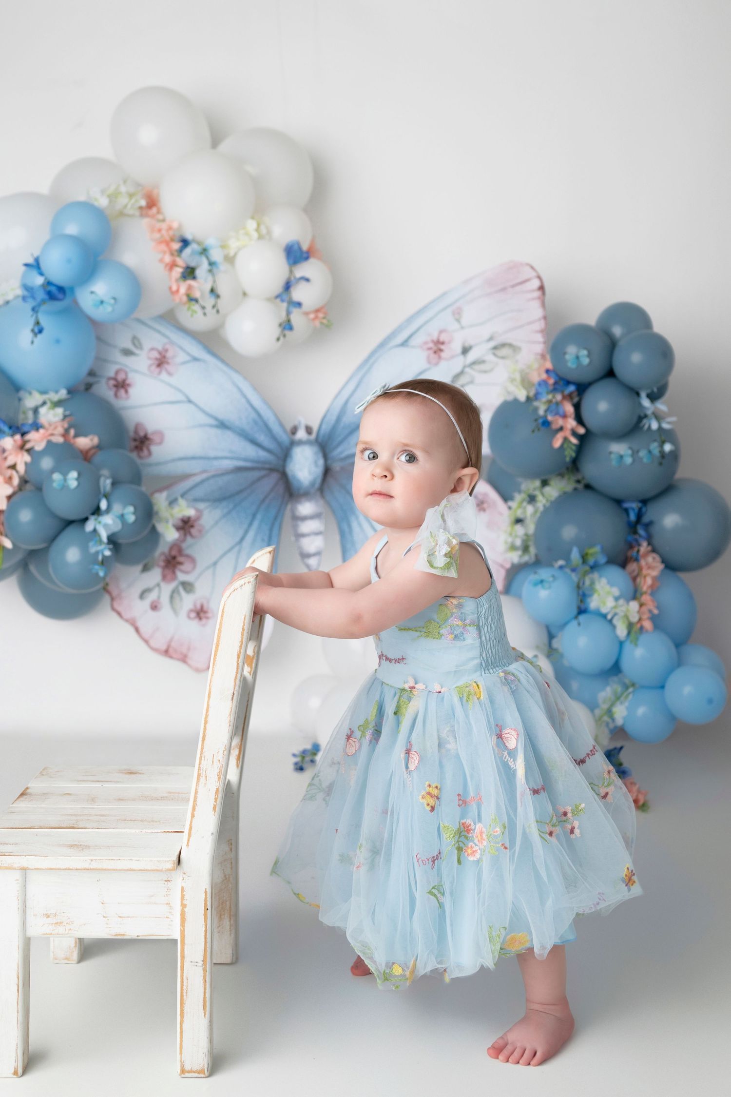 Baby in light blue tulle dress poses against white backdrop with blue balloon arch and floral decorations.