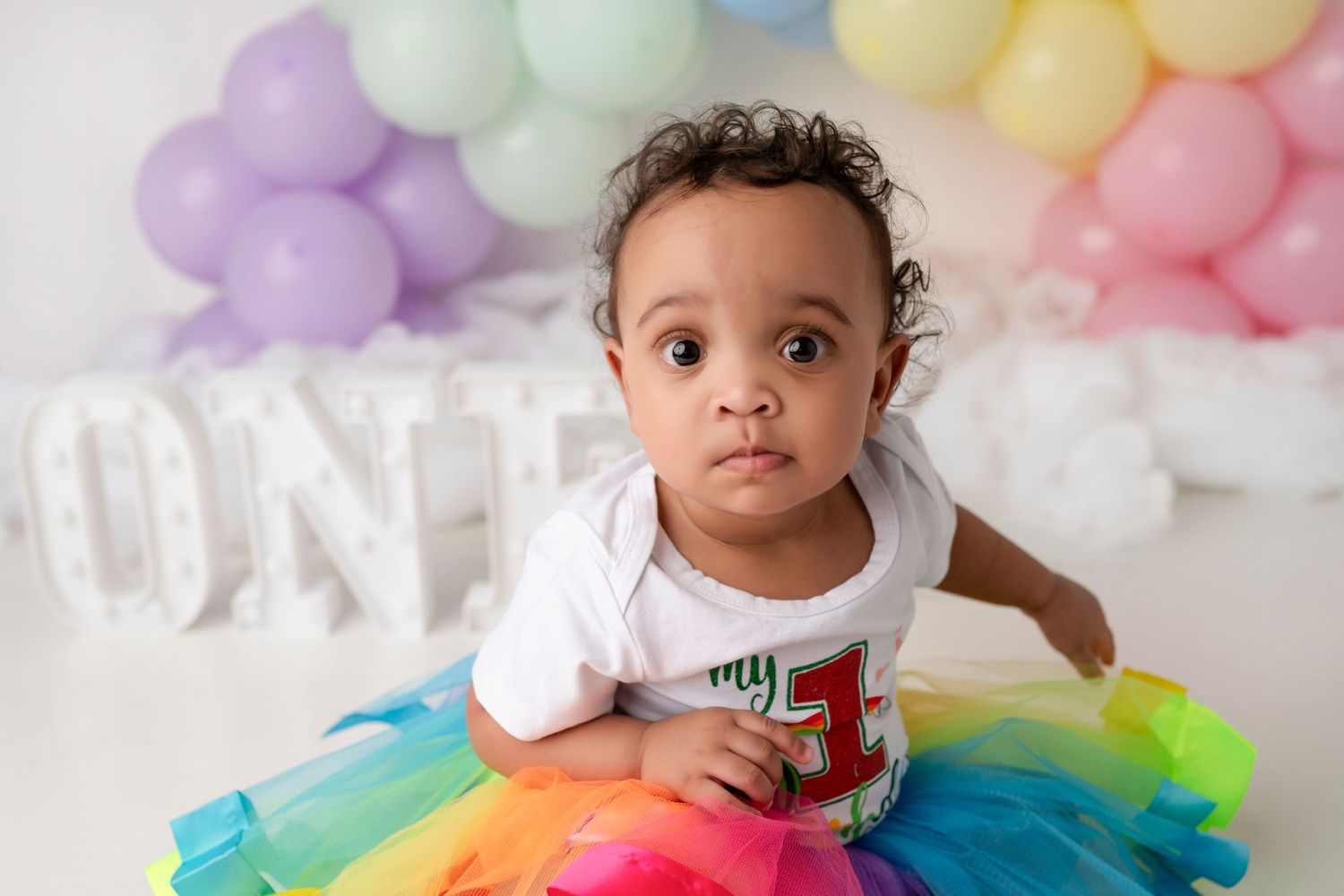 Baby in rainbow tutu sits in front of pastel balloon arch and white ONE letters for first birthday photoshoot.